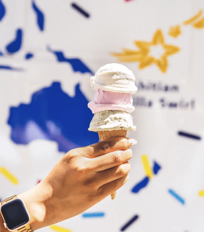 Hand with smartwatch holds triple-scoop ice cream in front of a wall with stars and blue shapes.
