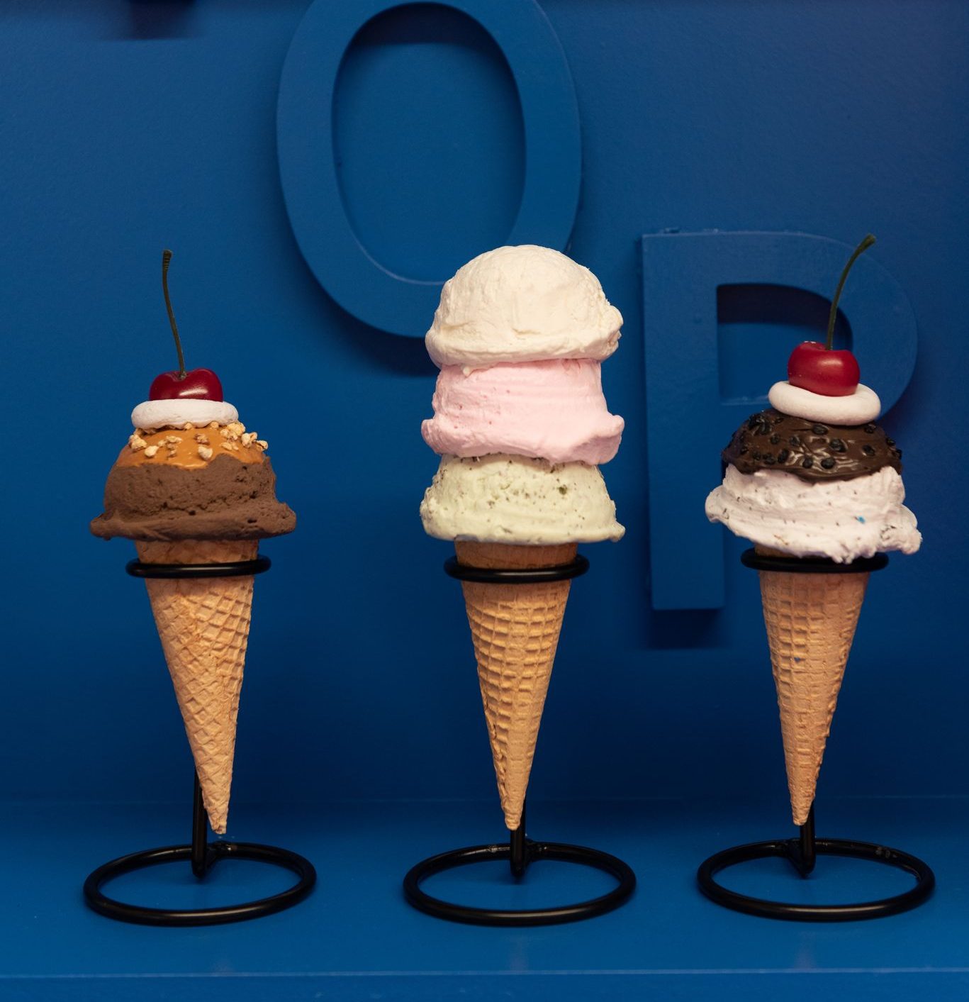 Three ice cream cones in holders against a blue background; each topped with a cherry, scoops vary.