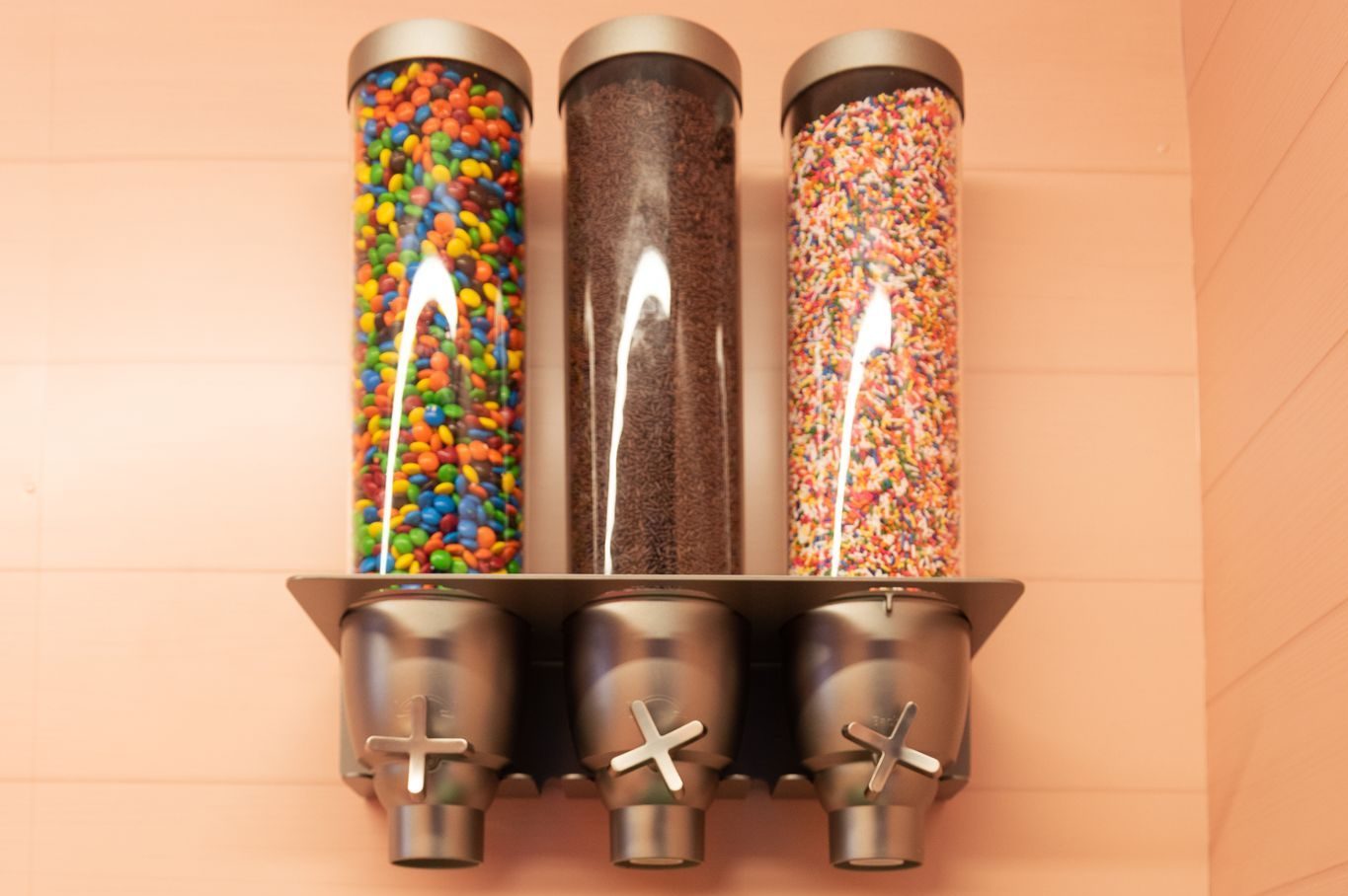 Three wall dispensers with candy-coated chocolates, chocolate sprinkles, and rainbow sprinkles.