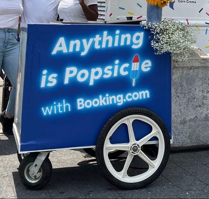 Blue popsicle cart with Booking.com branding, neon sign, popsicle icon, and two large white wheels.