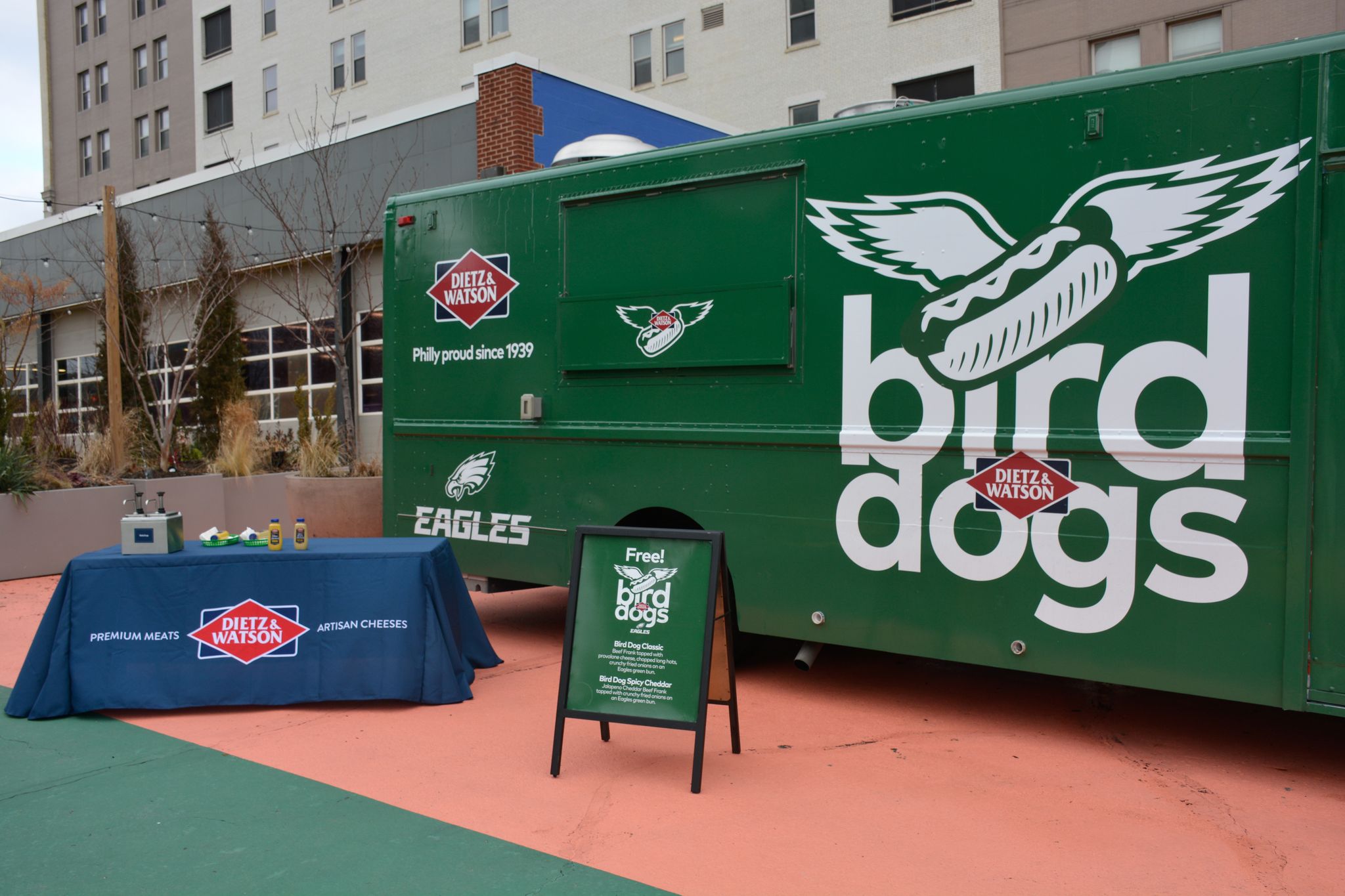 Green bird dog truck by Dietz & Watson table, sign offers free bird dogs, buildings behind.