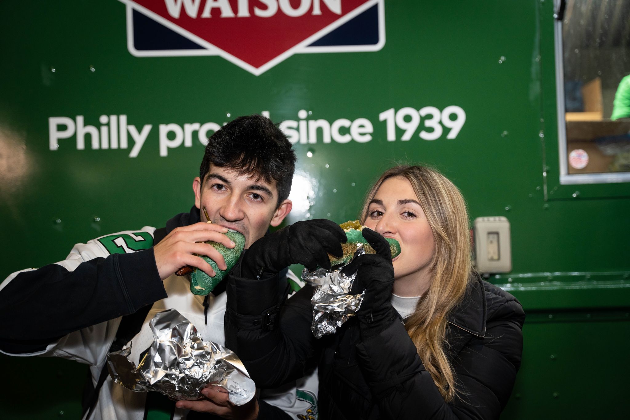 Two people in winter clothing eat green roll sandwiches by a green Bird Dog food truck.
