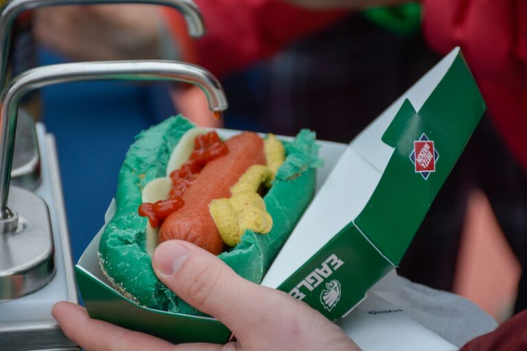 Person holding a green bun hot dog with ketchup and mustard near a condiment dispenser.