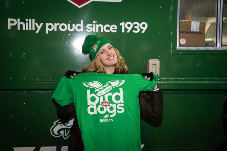 Woman in Eagles beanie holds green "bird dogs" T-shirt by wall saying Philly proud since 1939.