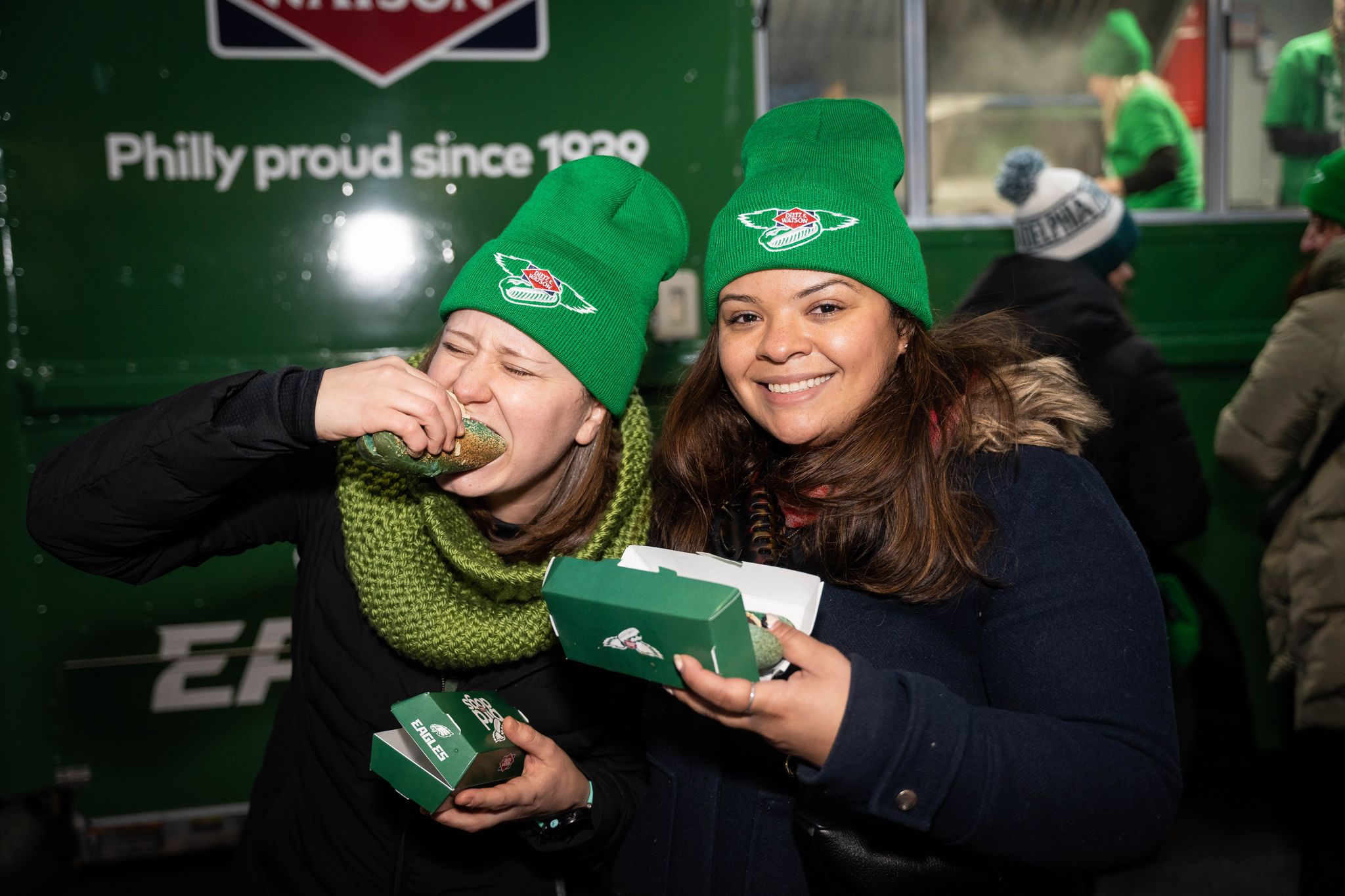 Two women in green hats hold food boxes and smile by a green Bird Dog Truck with people behind them.
