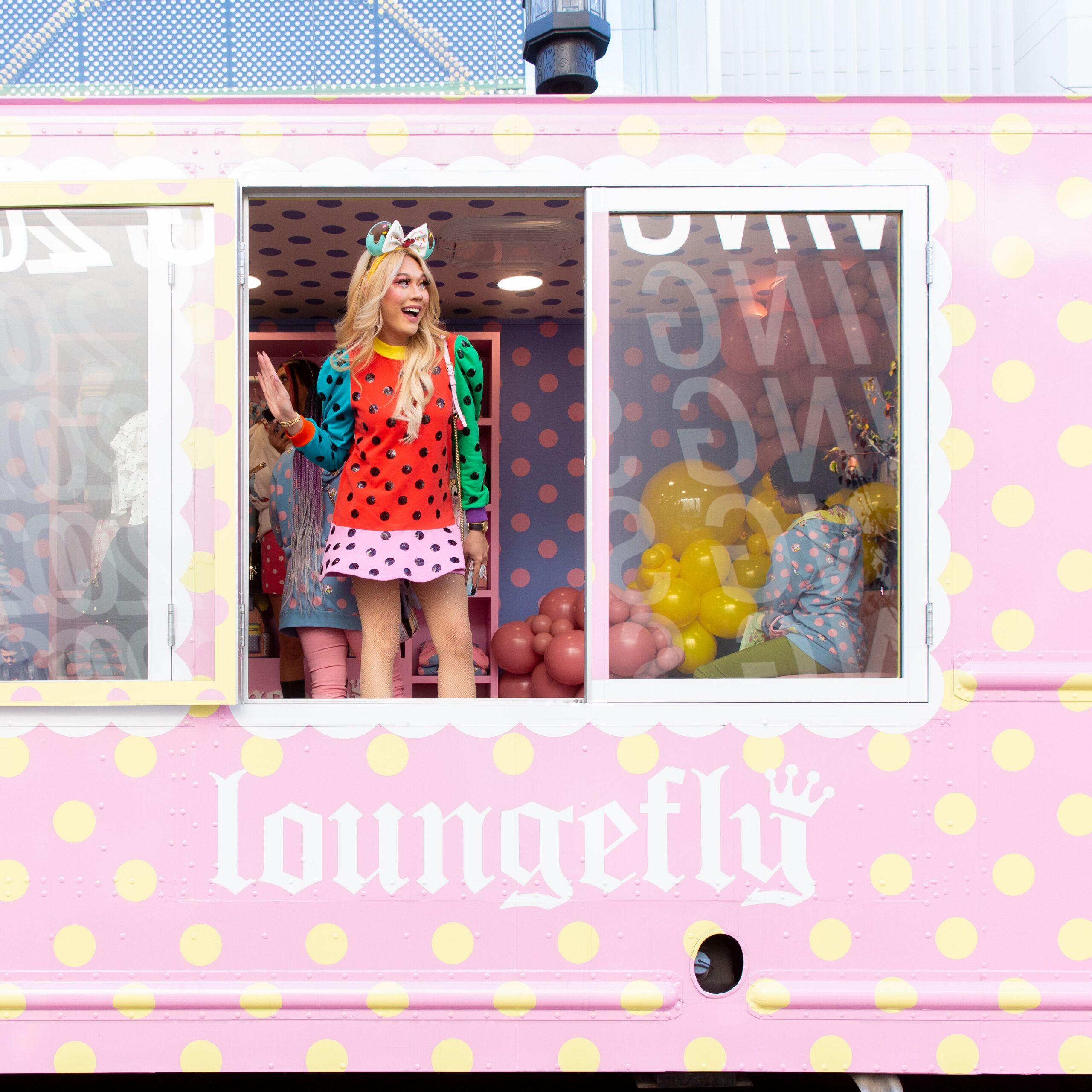 Woman in colorful clothes stands at a pink polka dot truck window with pastel balloons inside.