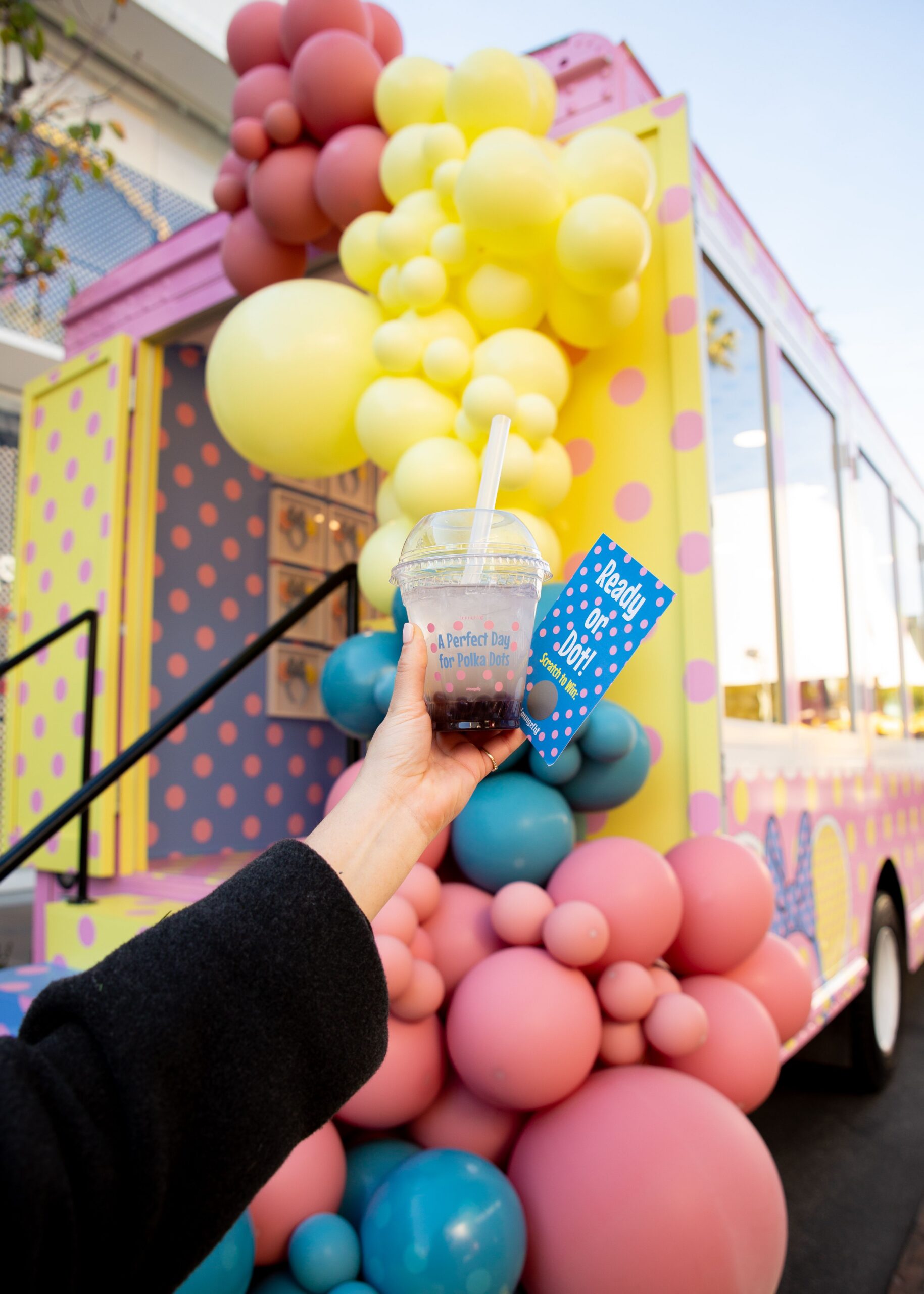 Hand holds a drink and blue card reading ready or not! by polka dot truck with big balloons.