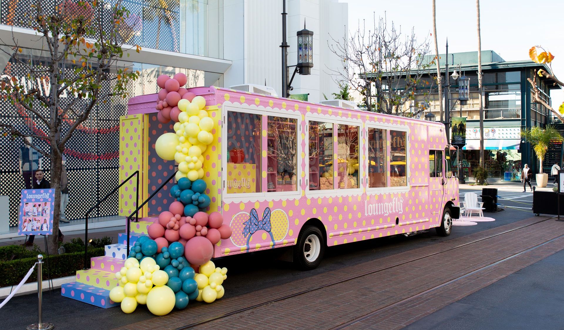 Pink and yellow polka dot pop-up truck with balloon clusters parked by a building and shops.