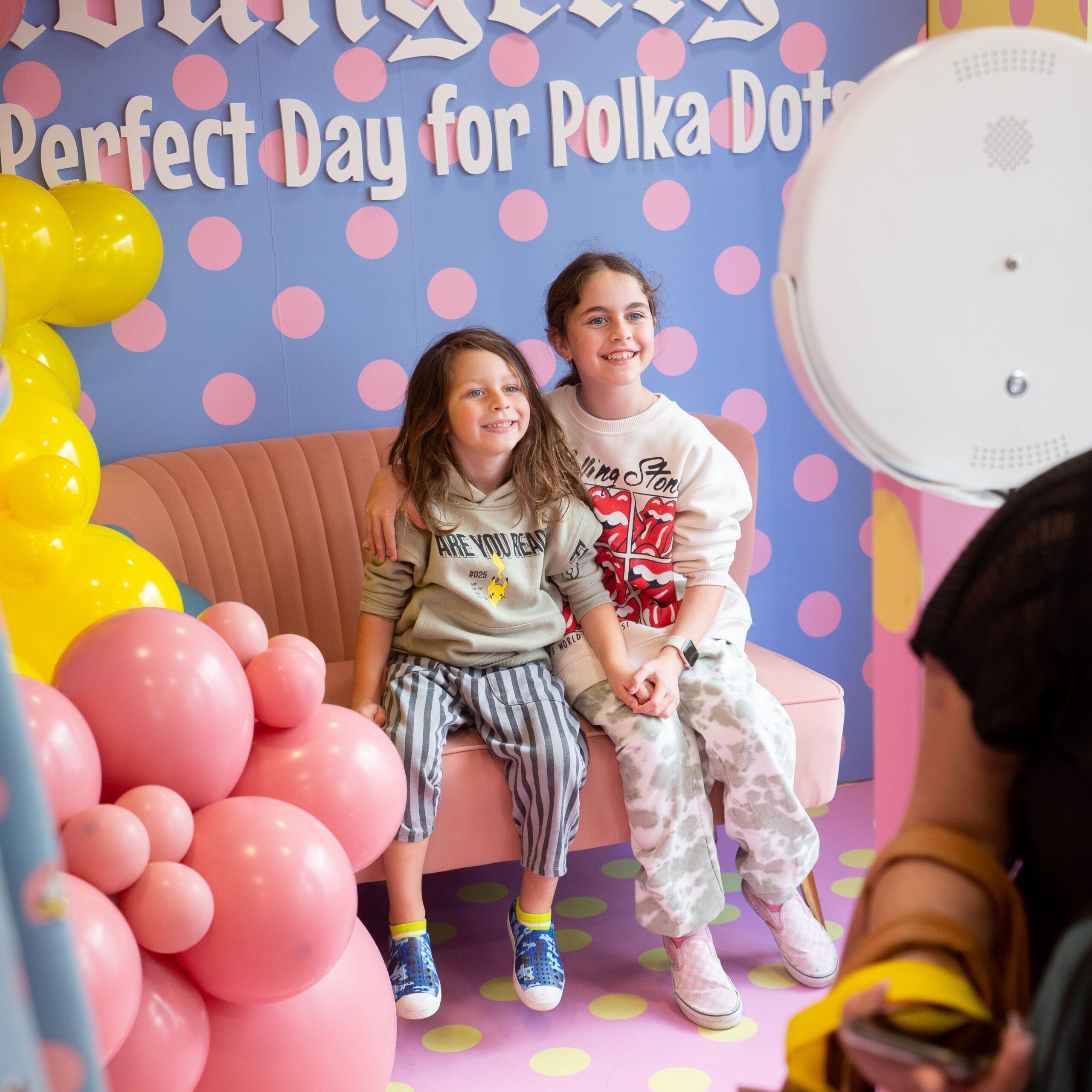 Two girls smiling on a pink couch with balloons, polka dot backdrop, and pop-up truck nearby.