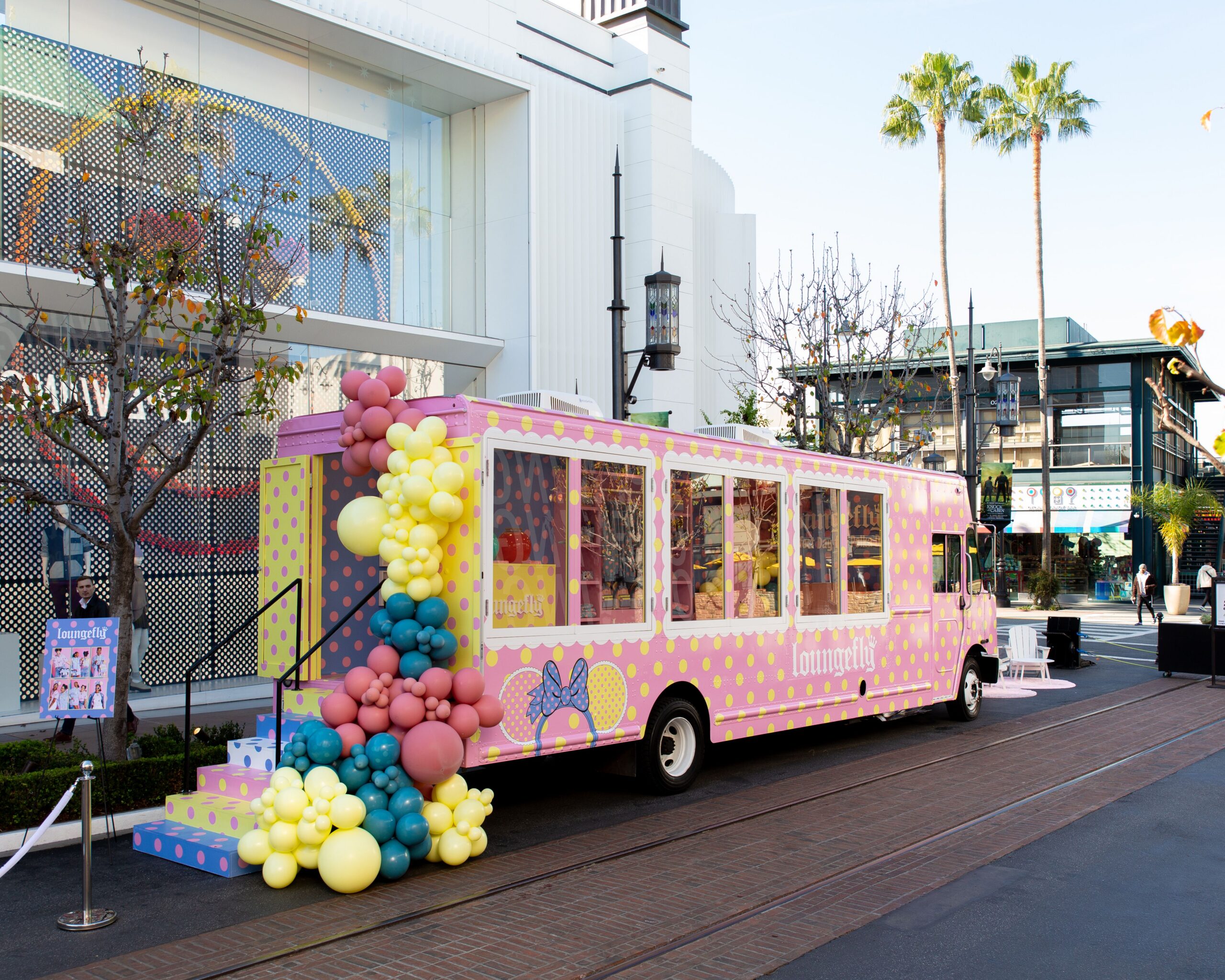 A pink and yellow polka dot truck with balloons is parked on a city street with palm trees.