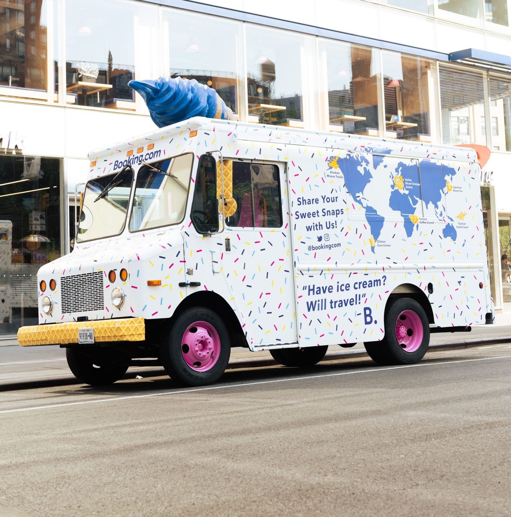 White booking.com ice cream truck with sprinkles and a blue cone parked by a glass building.