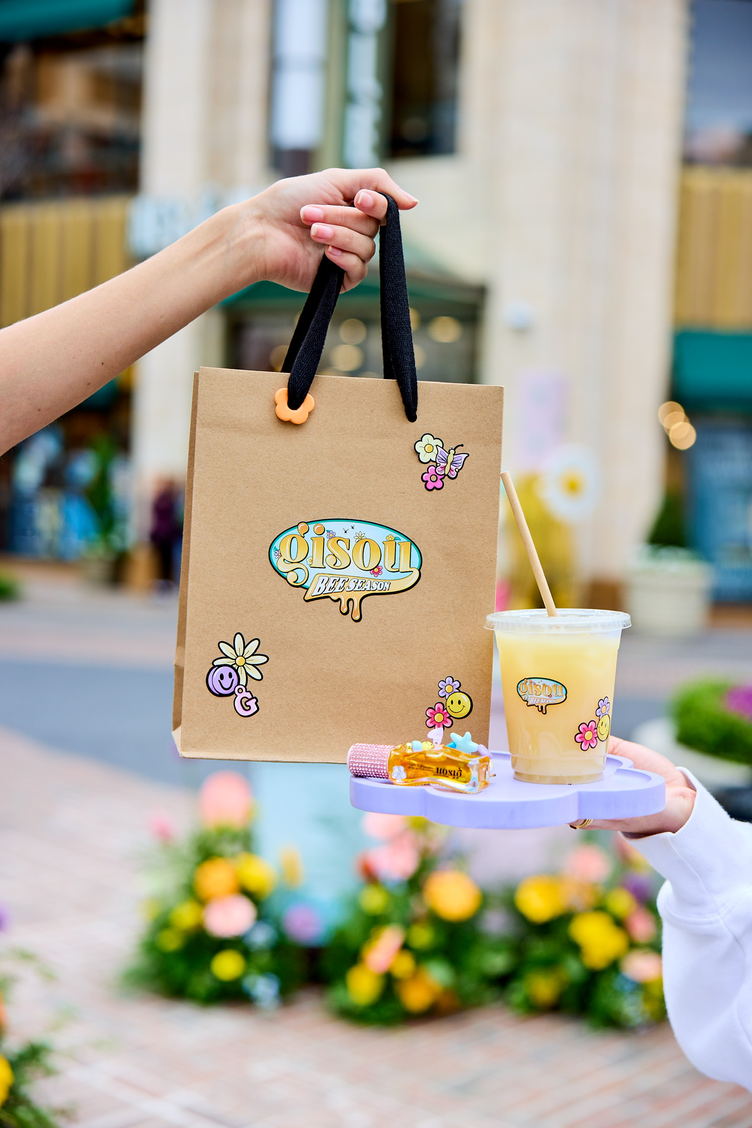 One hand holds a Gisou shopping bag; another holds a tray with a yellow drink and small bottle.