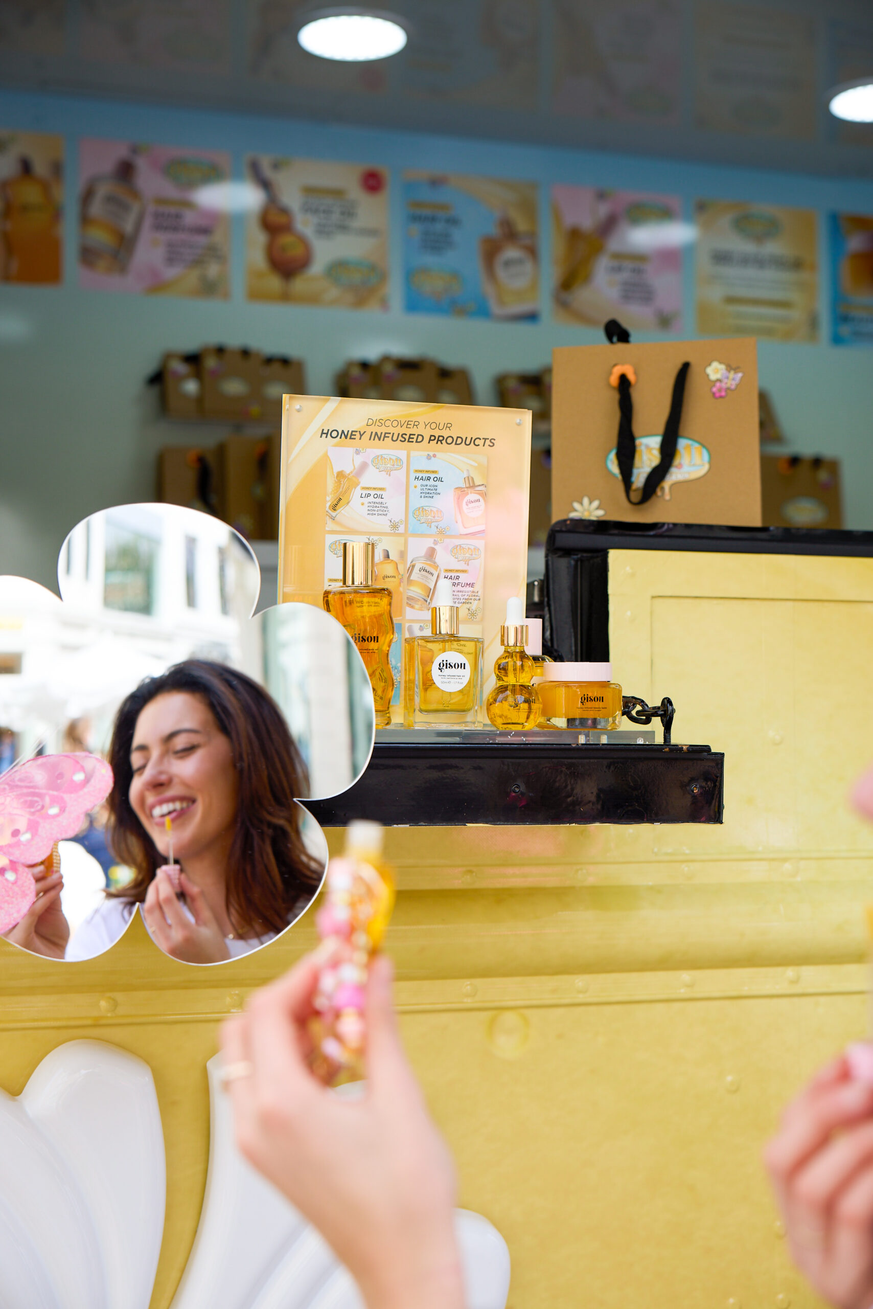 Woman applies flower-shaped lip balm; Gisou beauty products sit on a yellow counter behind her.