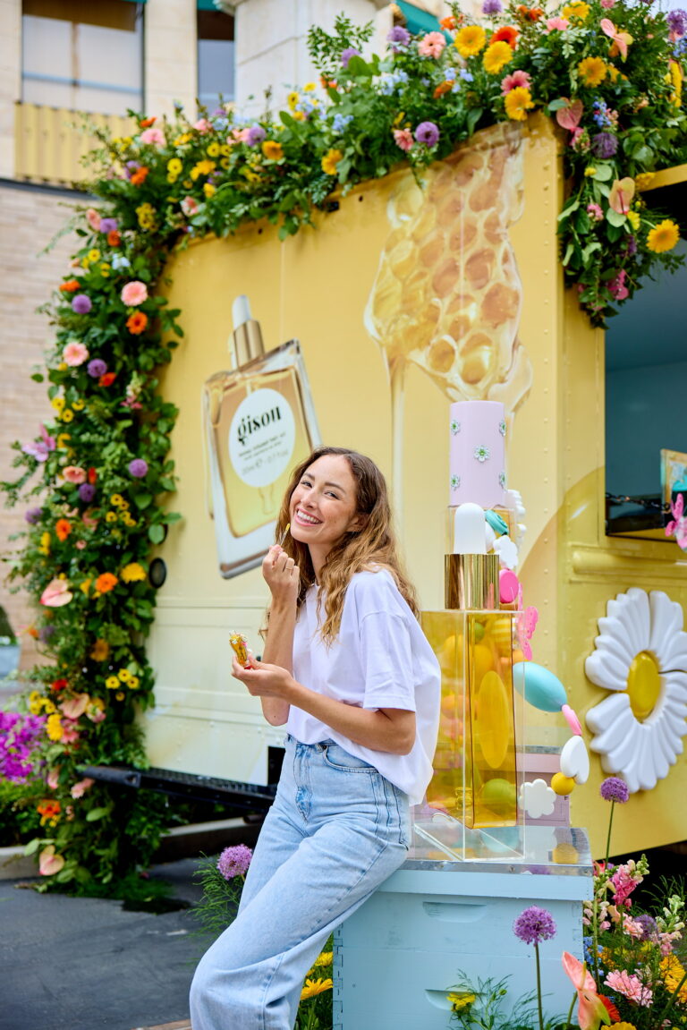 Woman in white t-shirt and jeans holds a treat by a flower-covered beauty truck and perfume display.