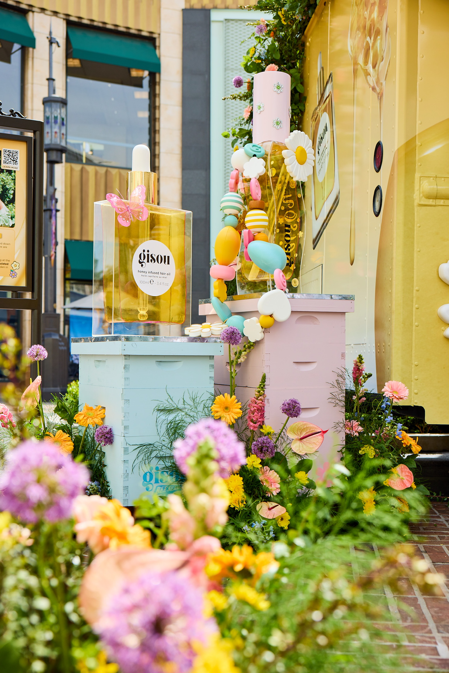 Pastel beehives with flowers, balloons, and large Gisou hair oil bottles by a yellow floral wall.