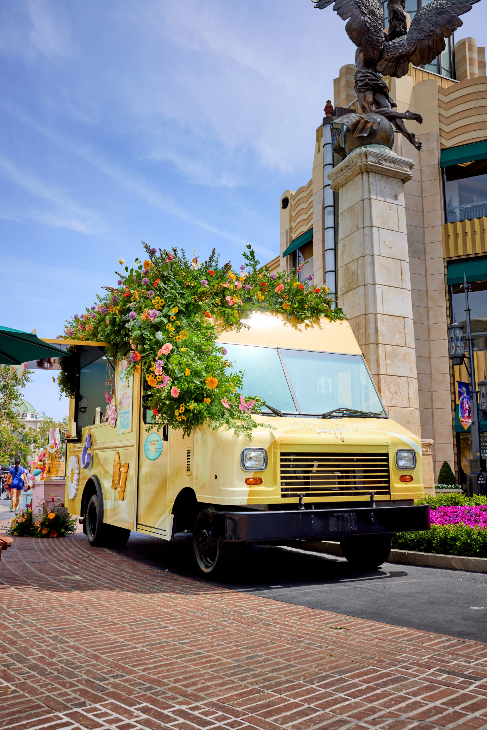 Yellow Gisou food truck with plants and flowers parked by tall building with winged statue.
