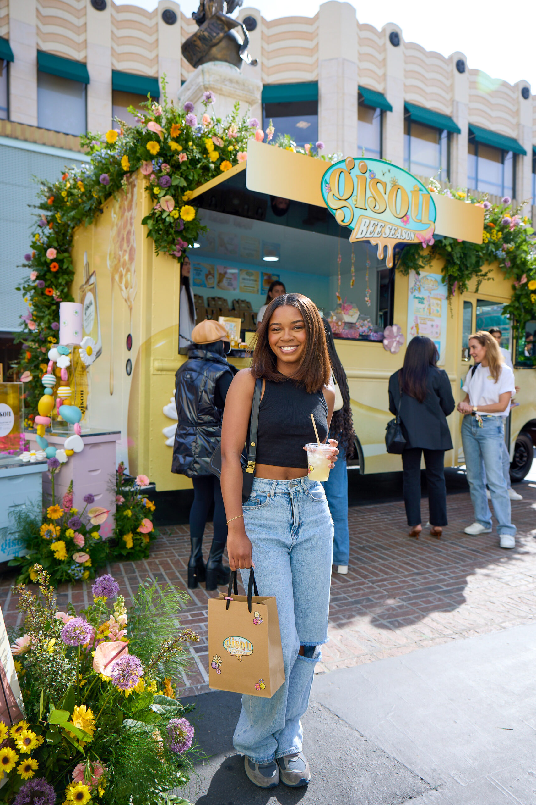 Woman with drink and shopping bag stands by Gisou “Bee-Yond” flower-decorated food truck.