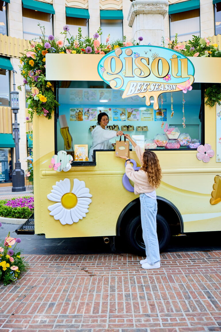 Woman gets a shopping bag from a vendor at a yellow flower-decorated Gisou truck outdoors.