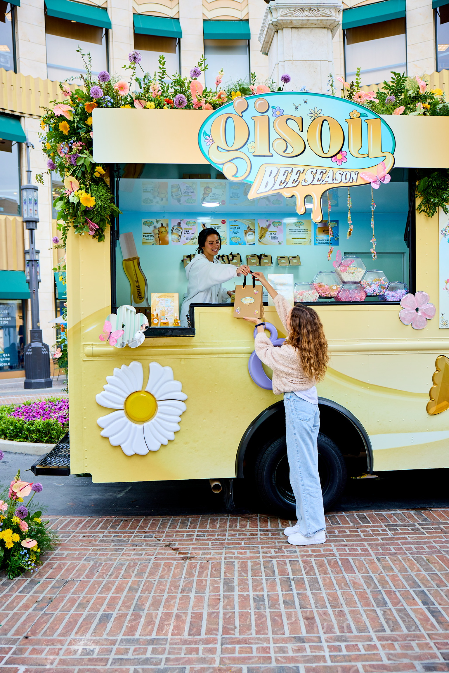 Woman gets a shopping bag from a vendor at a yellow flower-decorated Gisou truck outdoors.
