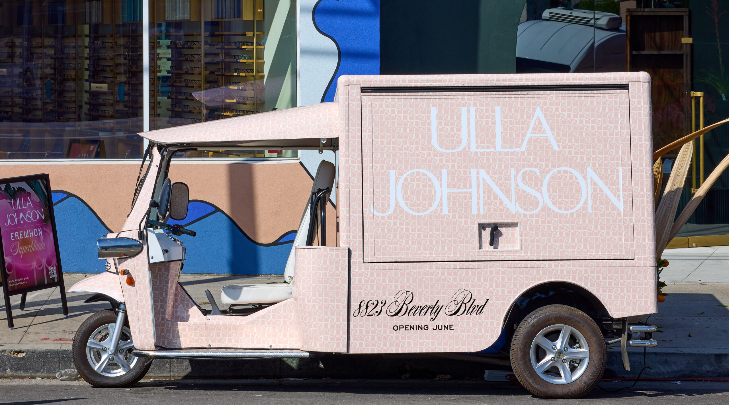 Pink tuk-tuk with “Ulla Johnson” and address parked by a building with murals and glass windows.
