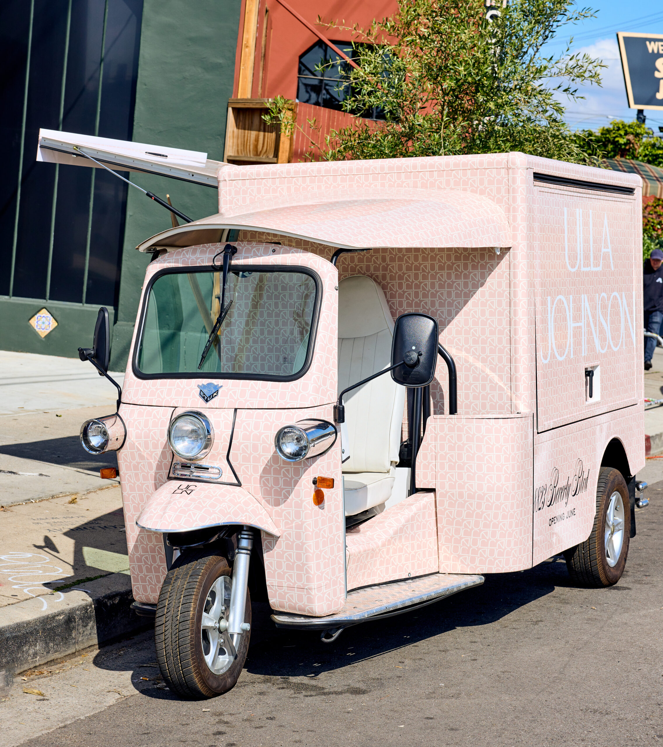 Pink three-wheeled Ulla Johnson food truck parked on street, serving window open, tree behind.