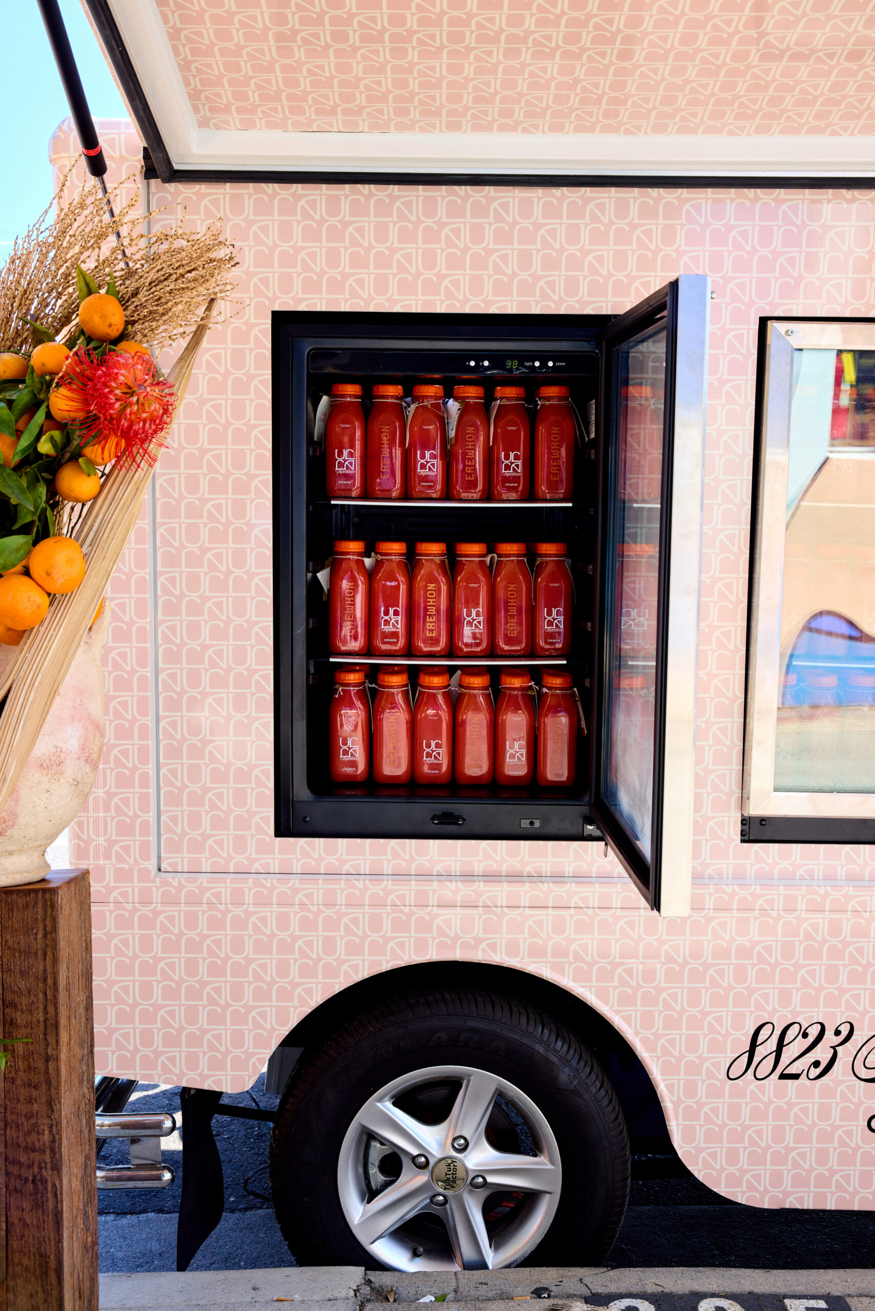 Food truck with open fridge of red juice bottles, flowers, citrus, and a visible car wheel.