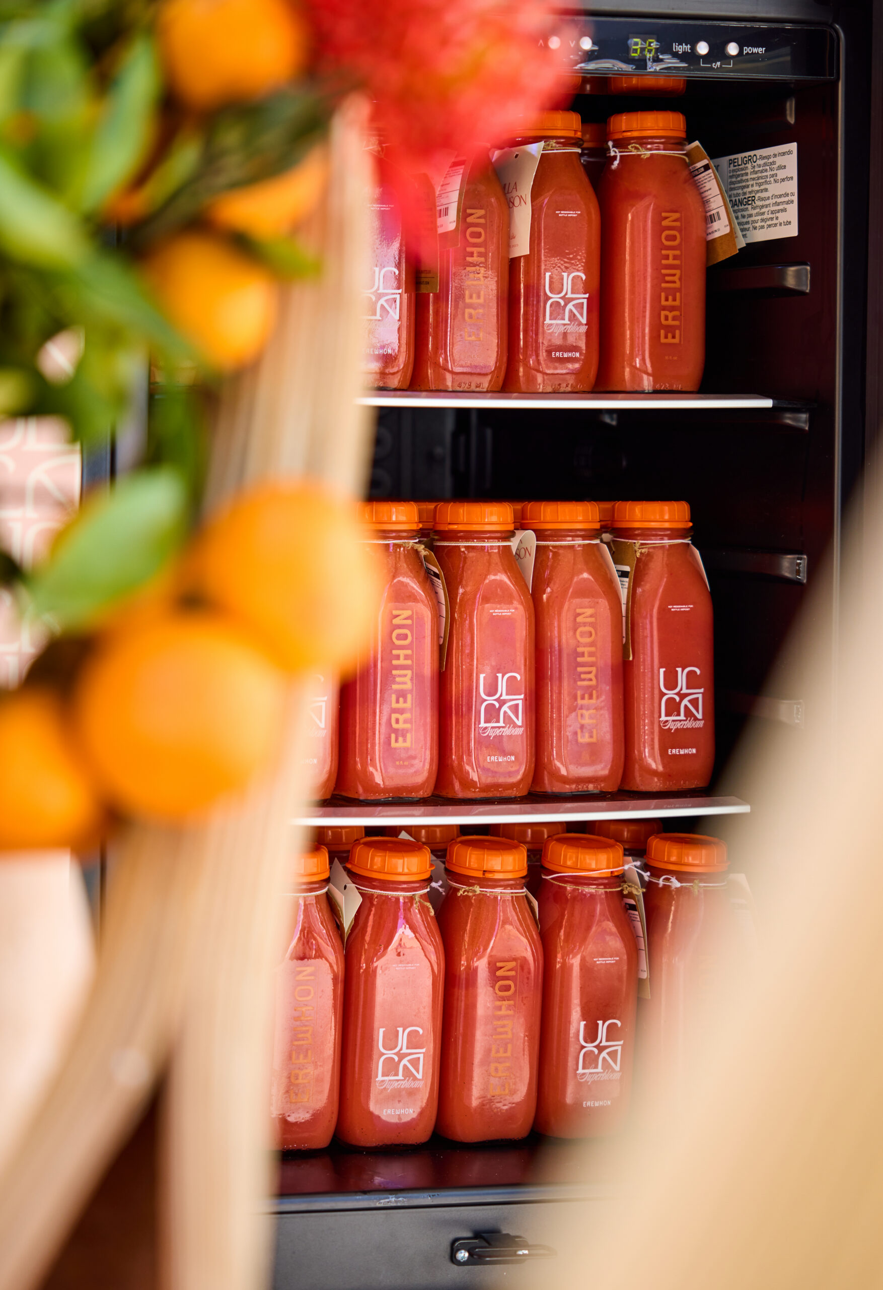 Erewhon juice bottles on refrigerator shelves with blurred oranges and flowers in front.