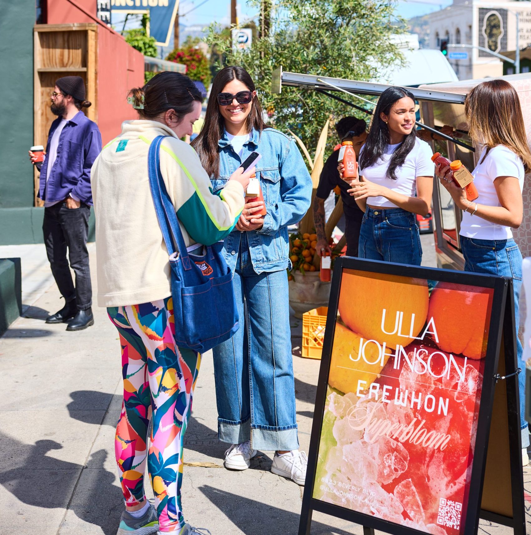 Women chat near an Ulla Johnson Erewhon Sunbloom sign, holding juice by a stand outdoors.