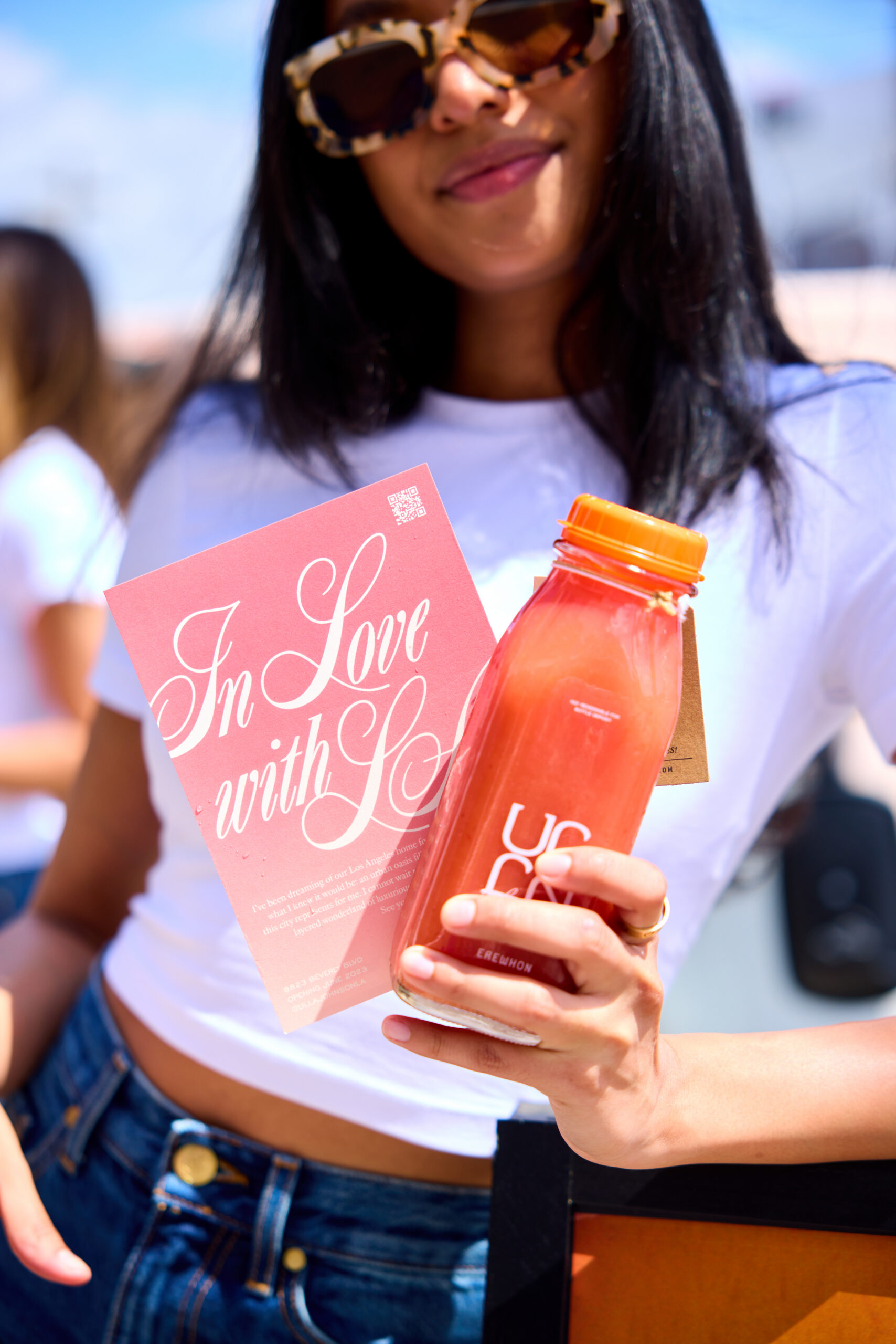 Woman in sunglasses and white t-shirt holds pink flyer and red juice bottle, smiling outdoors.