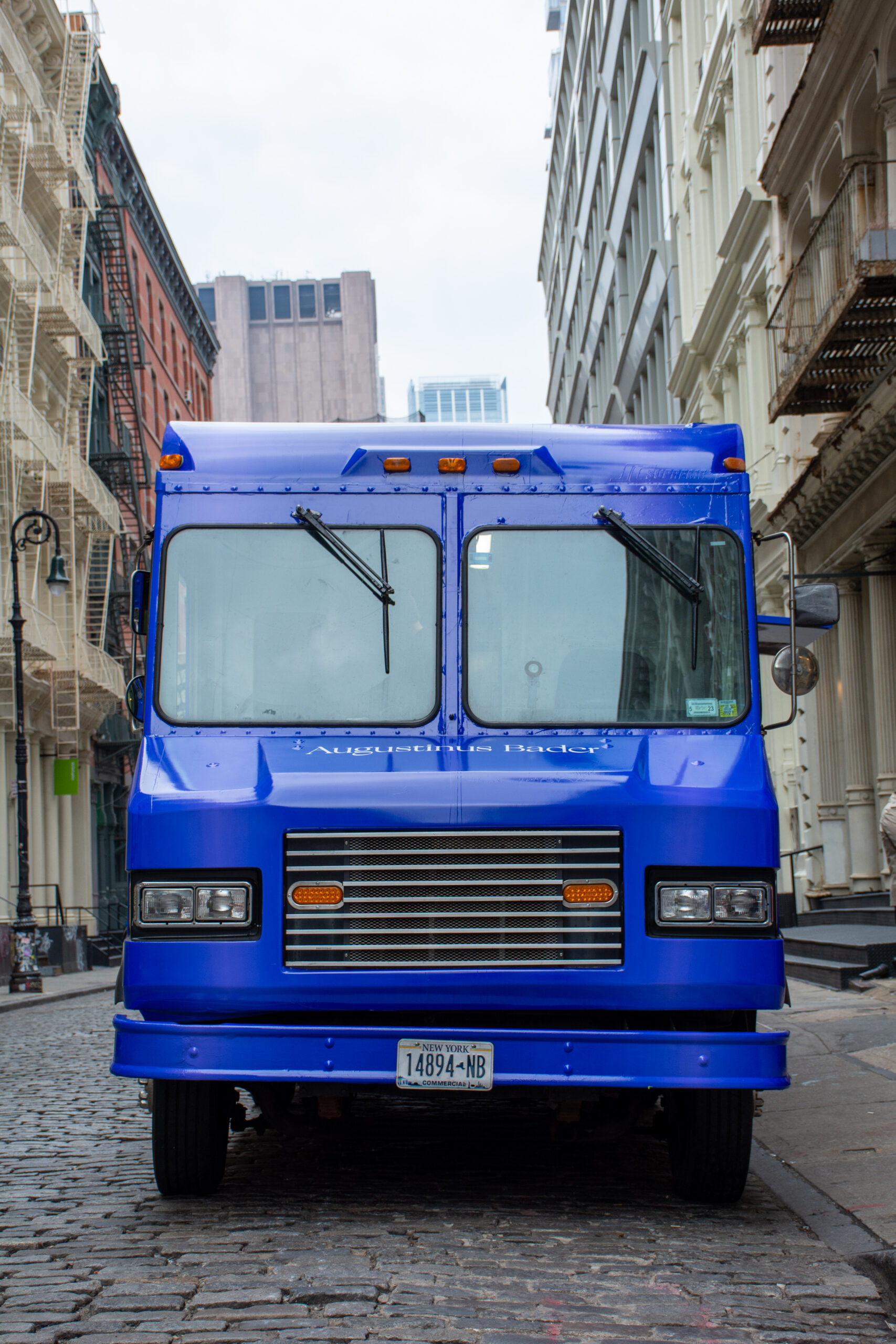 A blue Augustinus Bader delivery truck parked on a cobblestone street near tall buildings.