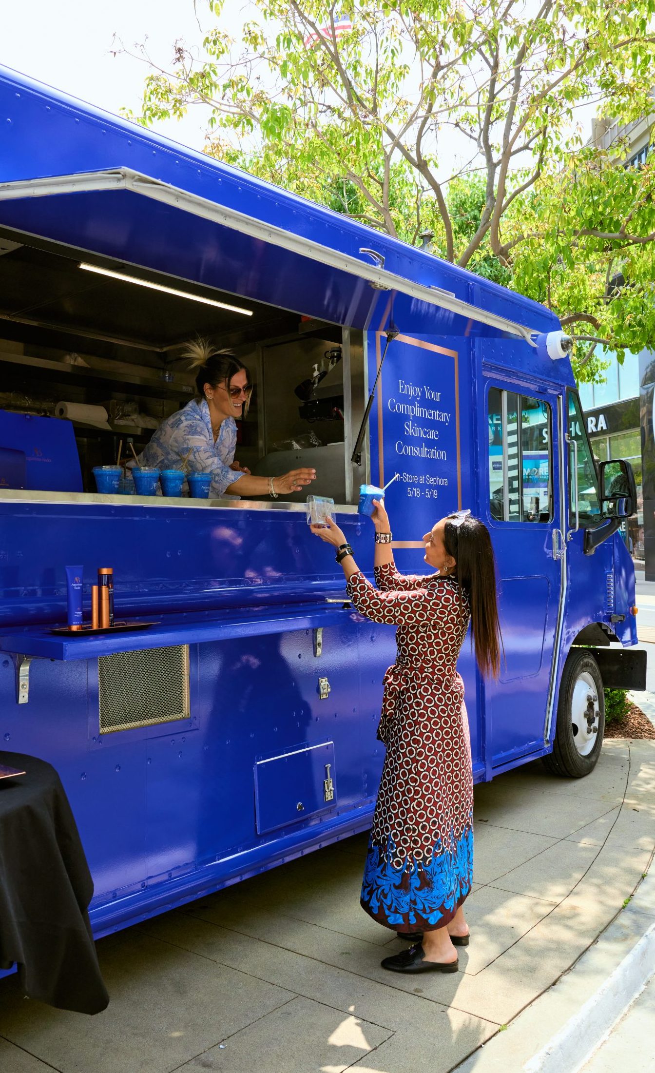 A woman gets an item from a worker at a blue Augustinus Bader food truck on a sunny sidewalk.