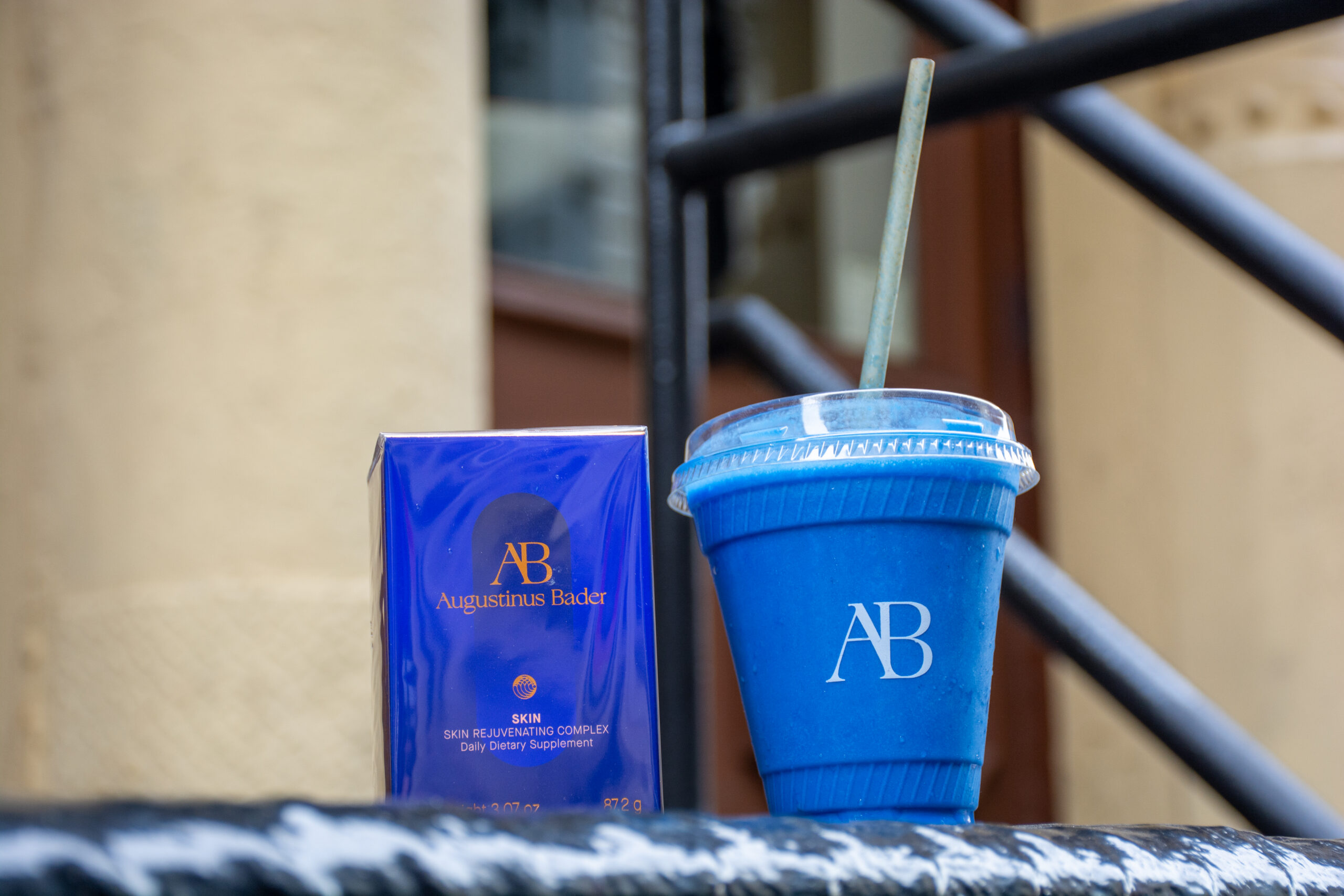 Blue smoothie cup with AB logo and straw beside Augustinus Bader supplement box on a stone ledge.