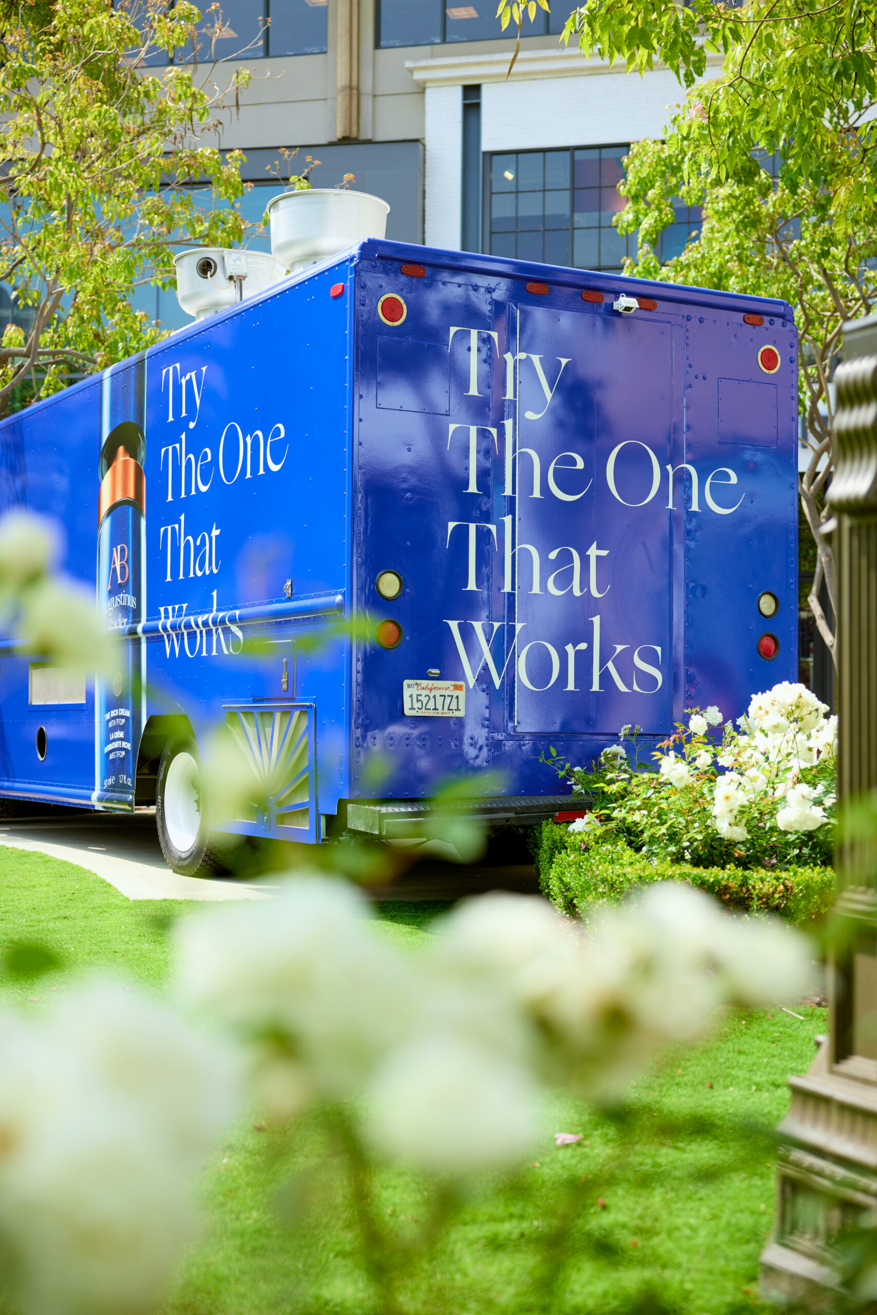 Augustinus Bader blue truck on grass shows “Try The One That Works”; trees and a building behind.