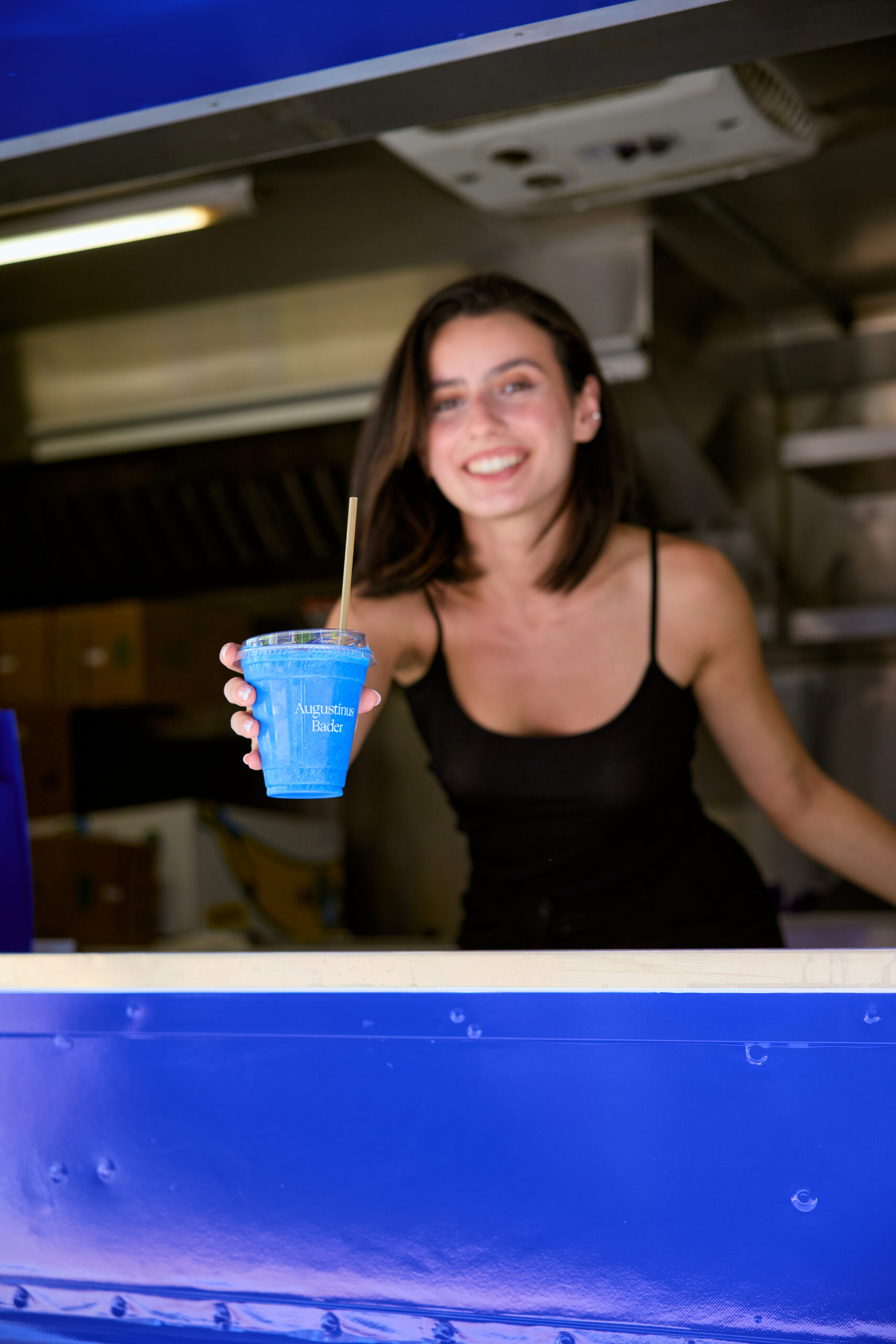 Woman in black tank top stands in food truck, offering a blue drink labeled Aquarius Bate.