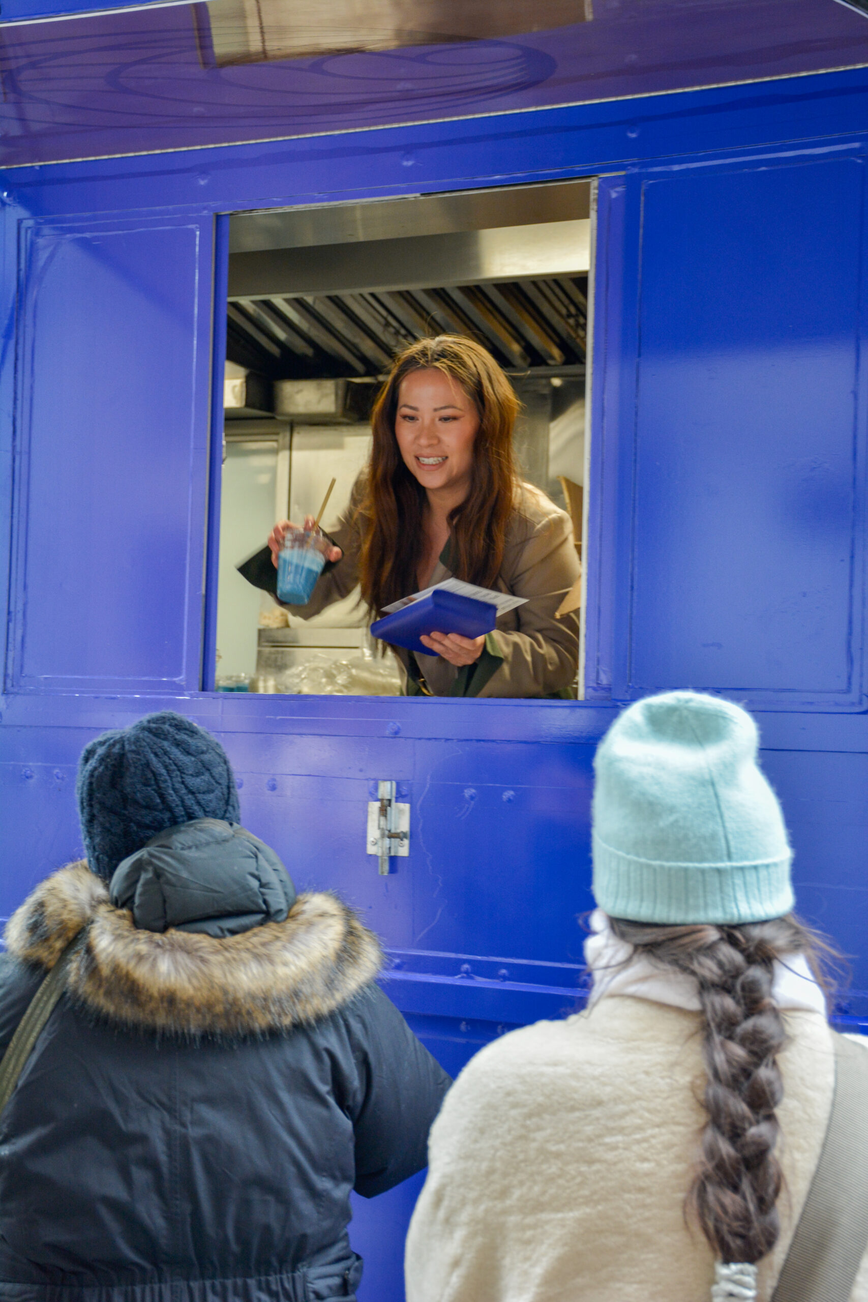 A woman in a blue food truck smiles, taking an order from two people in winter coats.