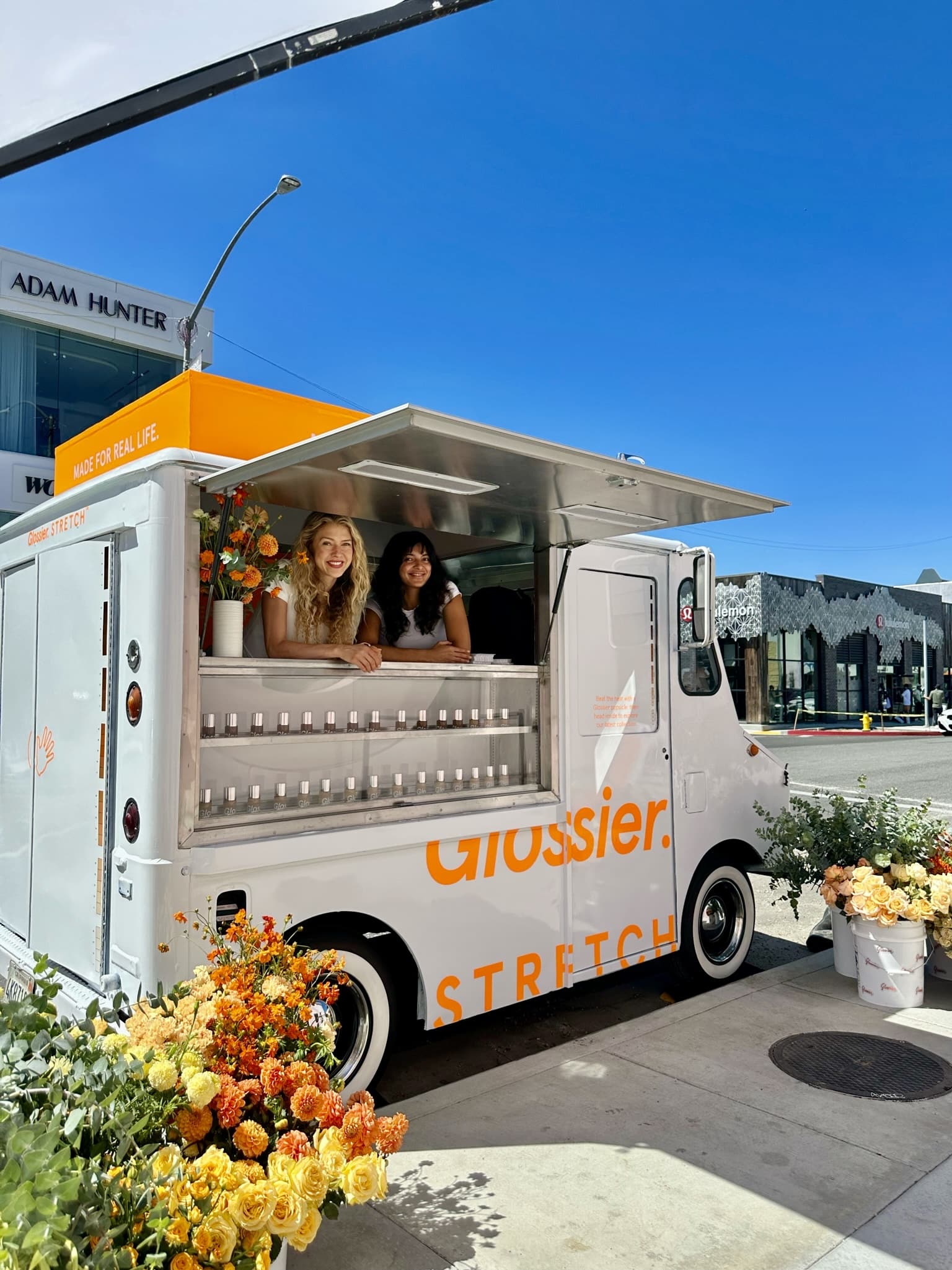 Two women smile in a white Glossier truck with &ldquo;STRETCH&rdquo; branding and flower displays.
