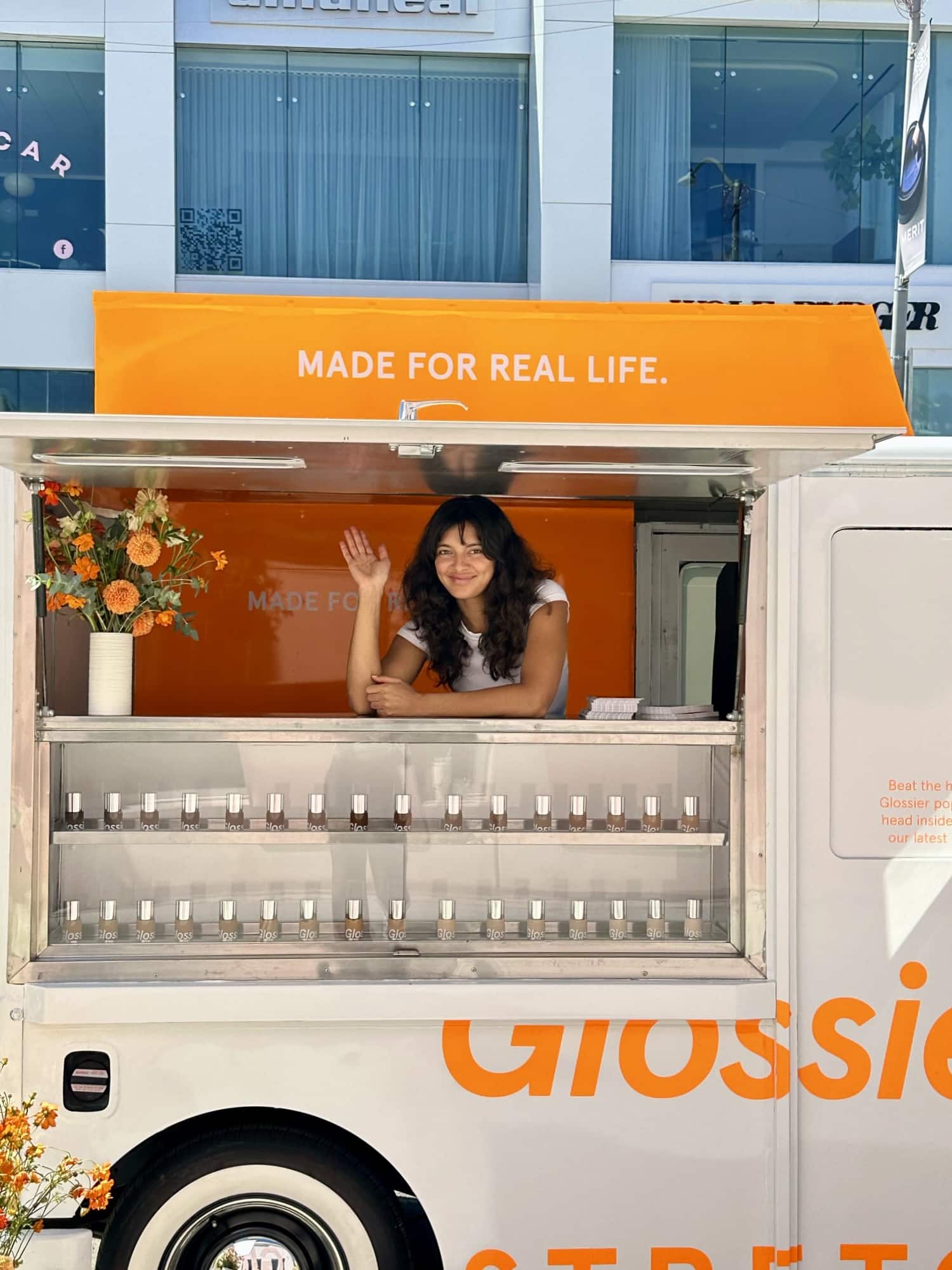 Woman waves and smiles from a Glossier truck with an orange sign; cosmetics and flowers nearby.