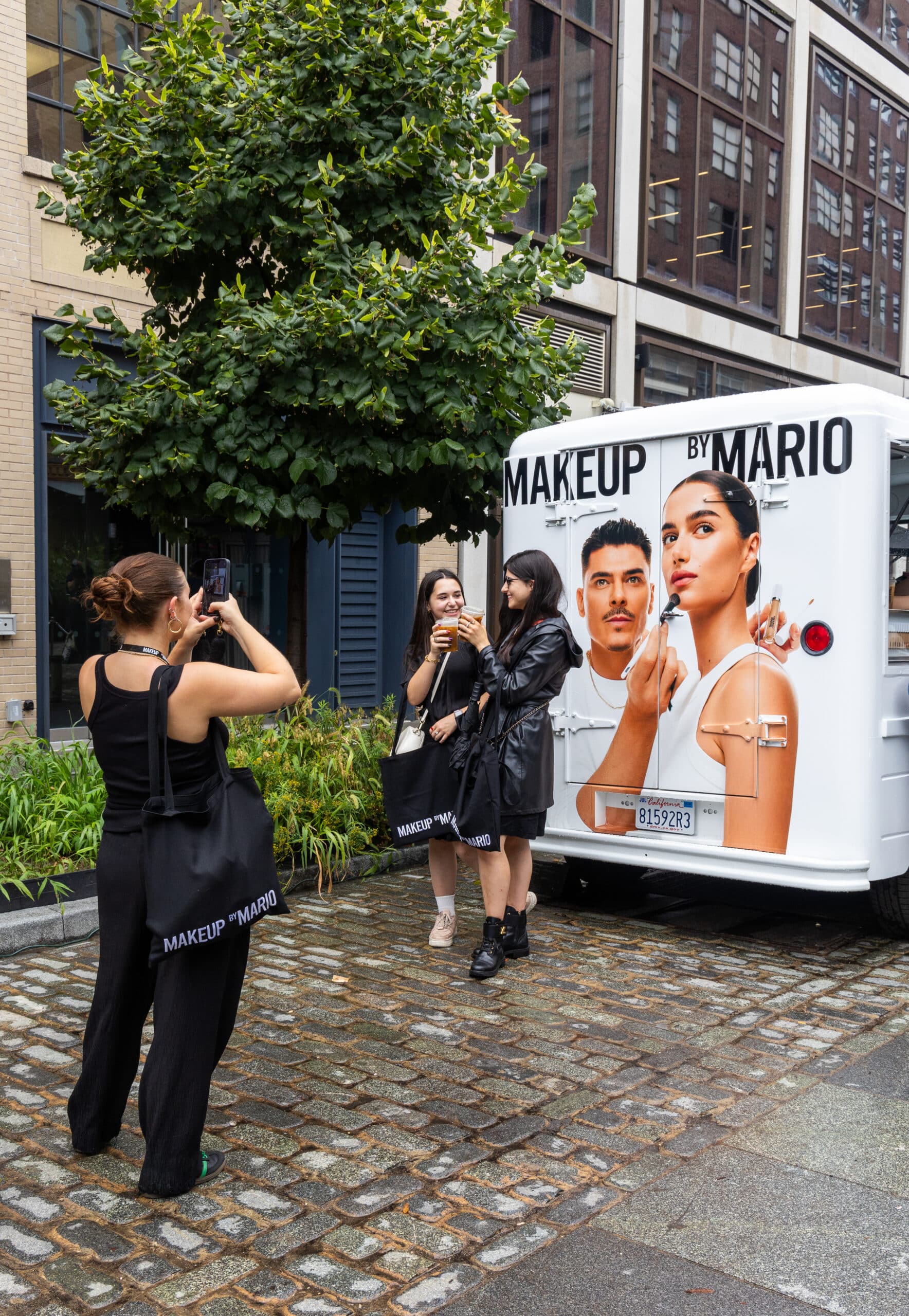 Three women with branded tote bags pose by a Makeup by Mario truck on a cobblestone street.
