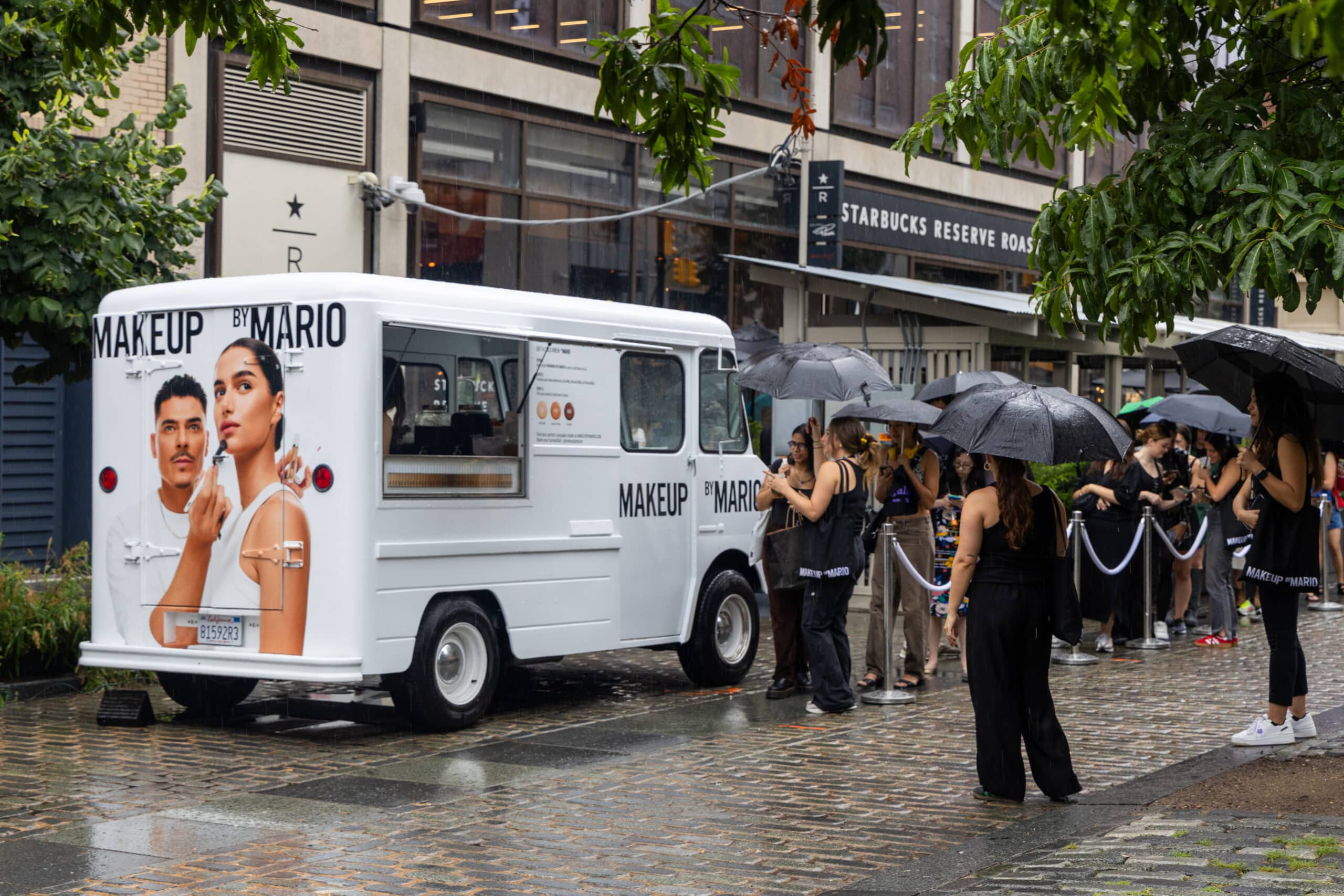 White MAKEUP BY MARIO van parked on rainy street; people with umbrellas line up nearby.