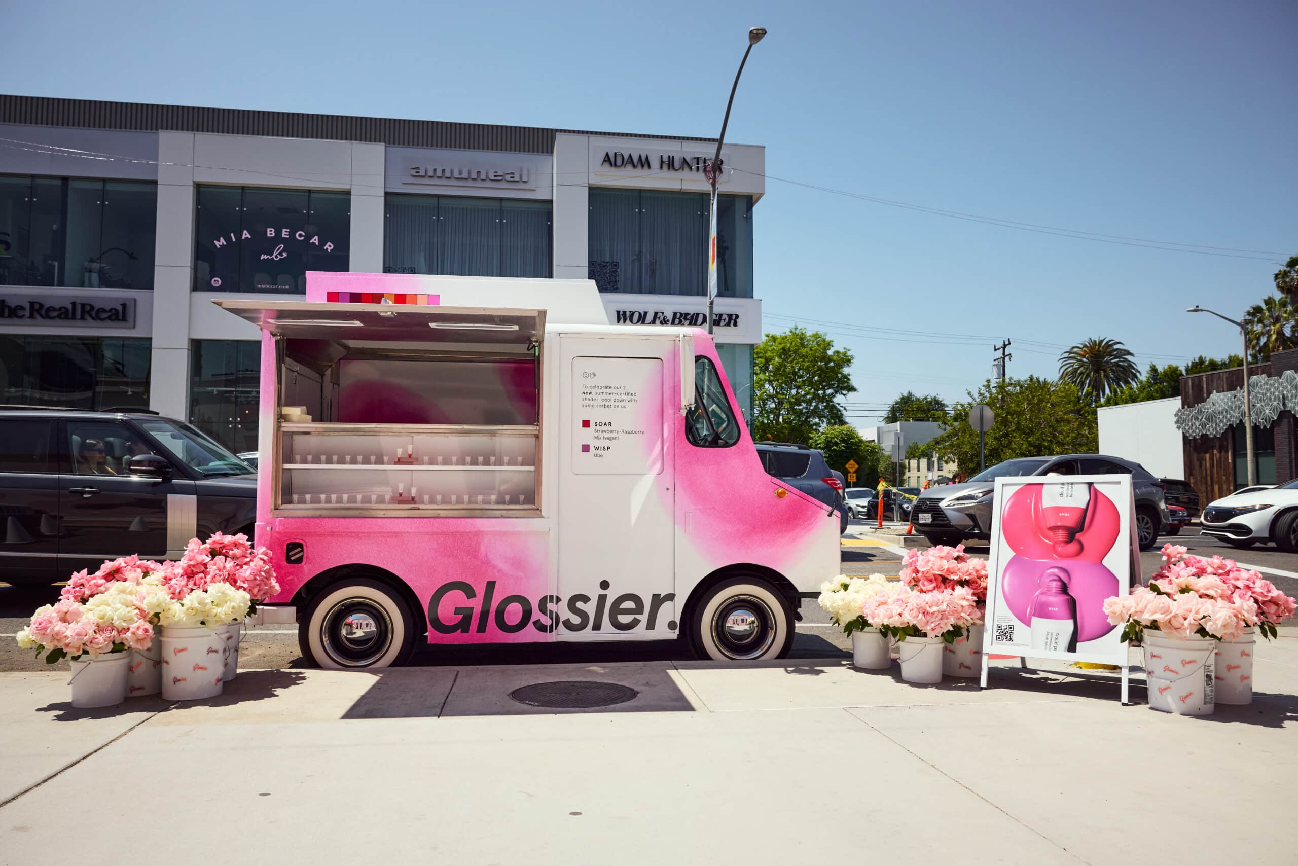 Glossier pink and white truck on sidewalk with flower buckets, product display, and buildings.