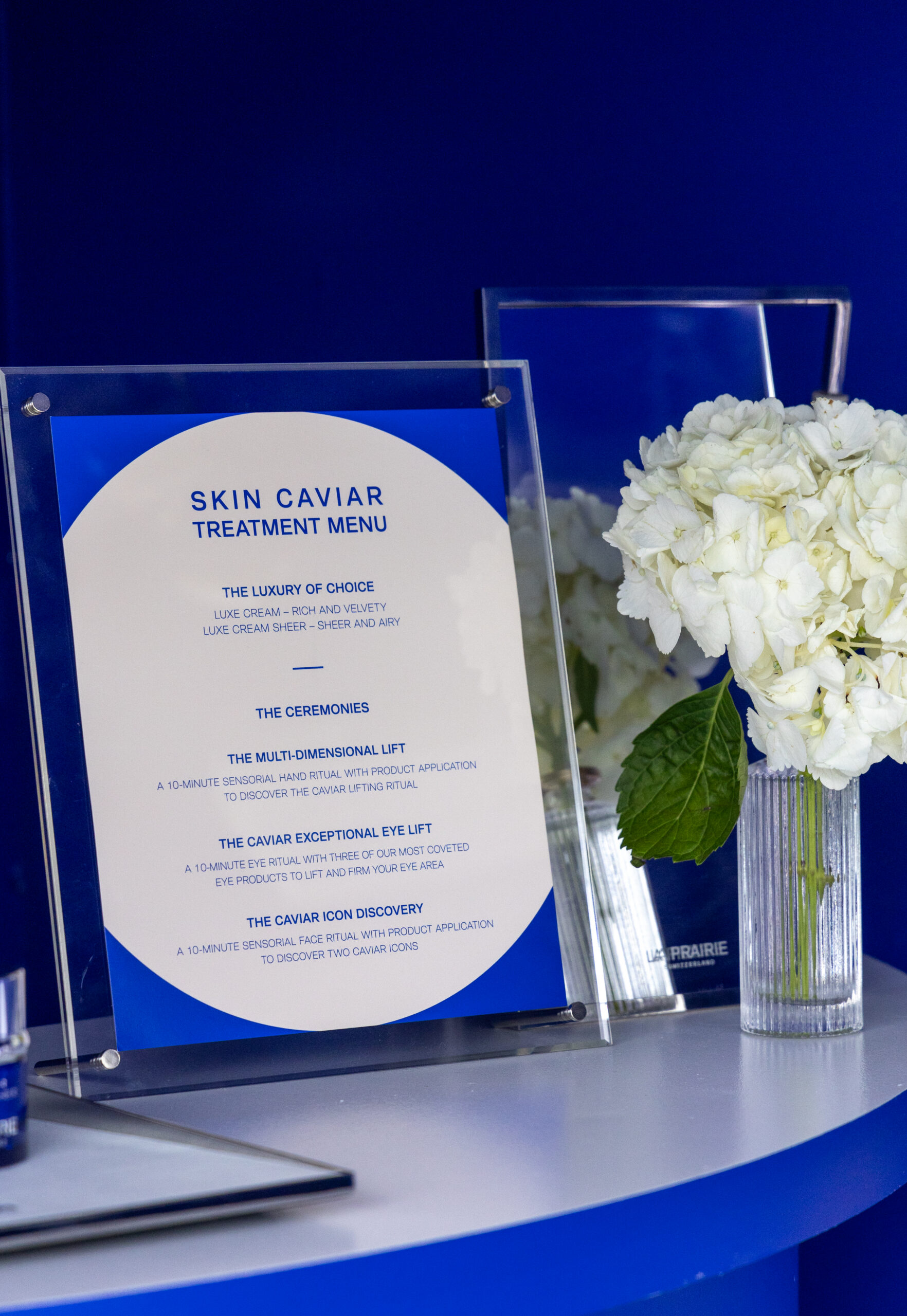 Framed Skin Caviar Treatment Menu on blue table beside white hydrangeas and a mirror.