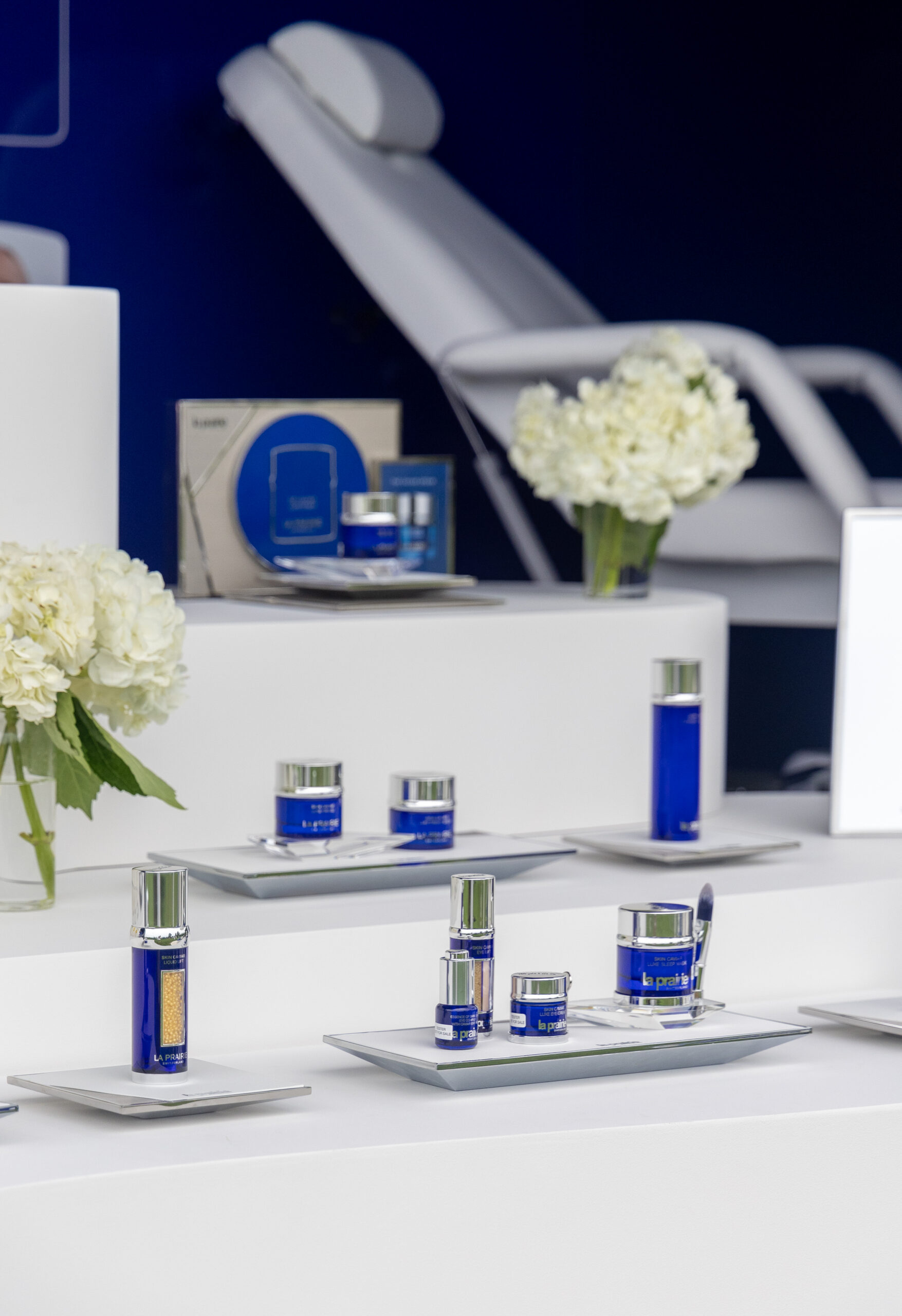 Blue and silver skincare products on white platforms with hydrangeas; treatment chair behind.