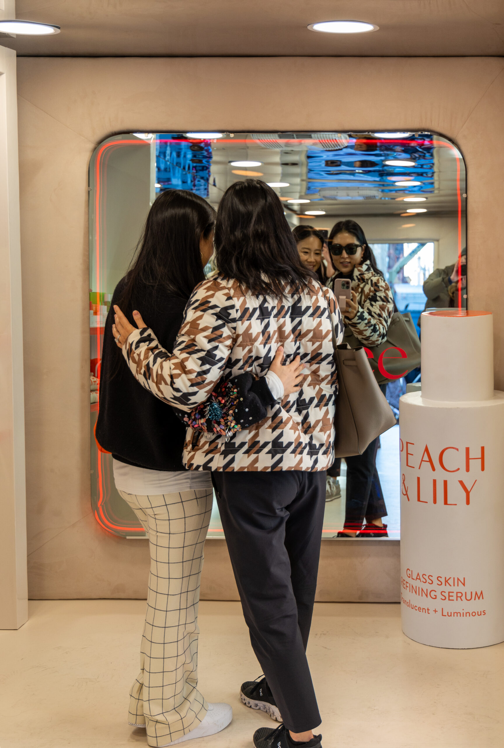Two women with arms around each other face a mirror near a Peach & Lily display in a store.