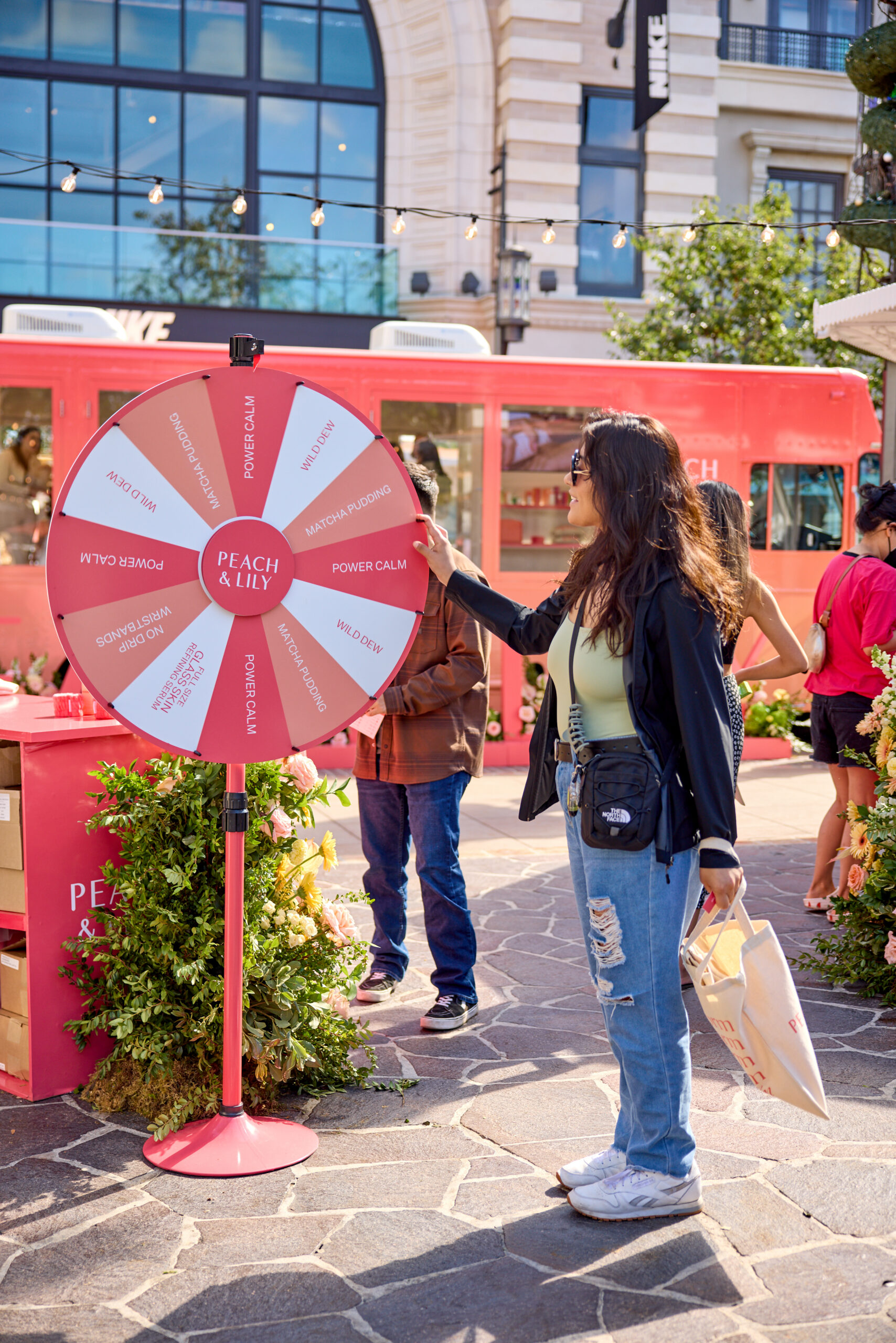 Woman in sunglasses spins a skincare prize wheel at an outdoor Peach & Lily event by a pop-up bus.