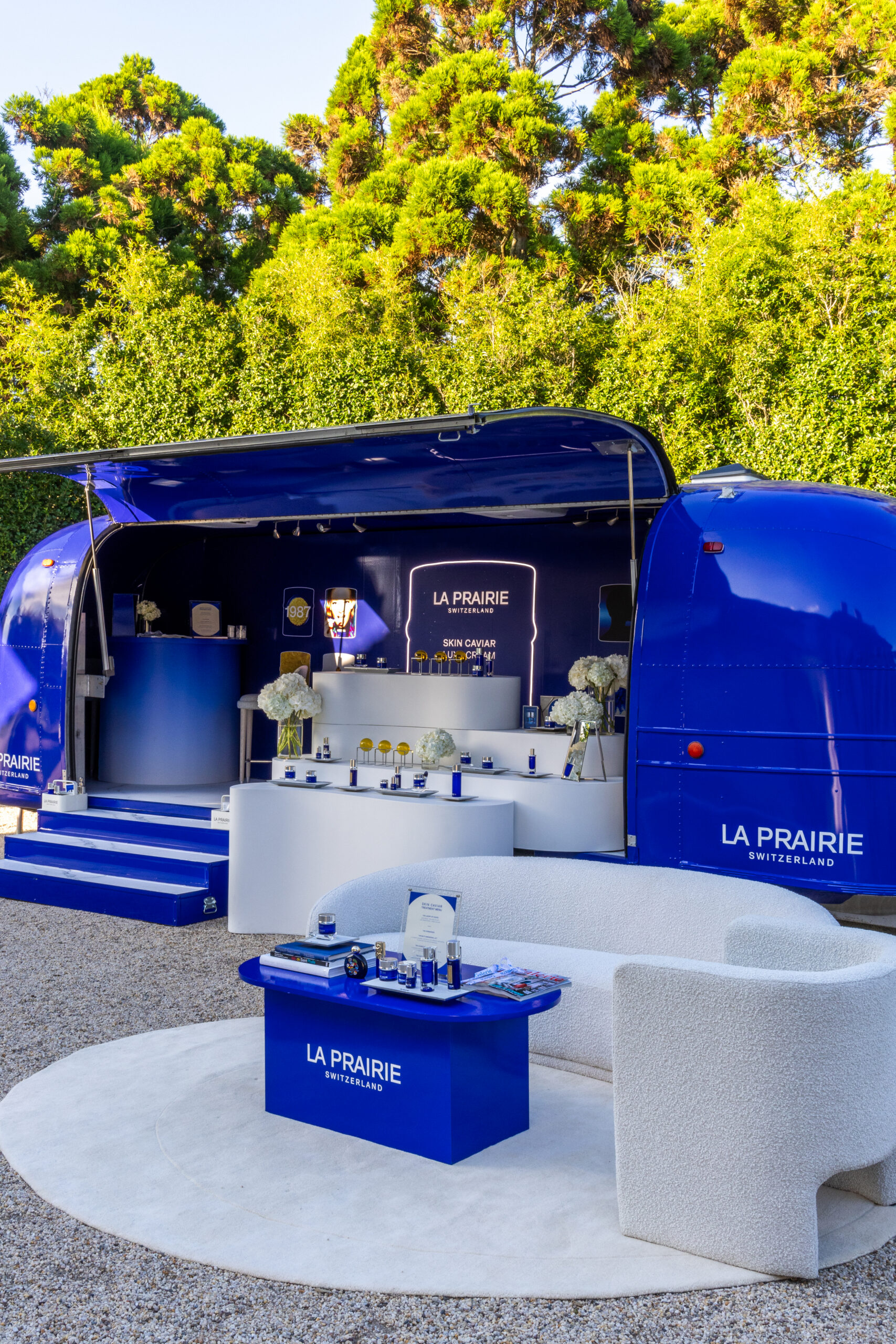 La Prairie blue pop up trailer displays skincare, with a white sofa, table, and trees behind.