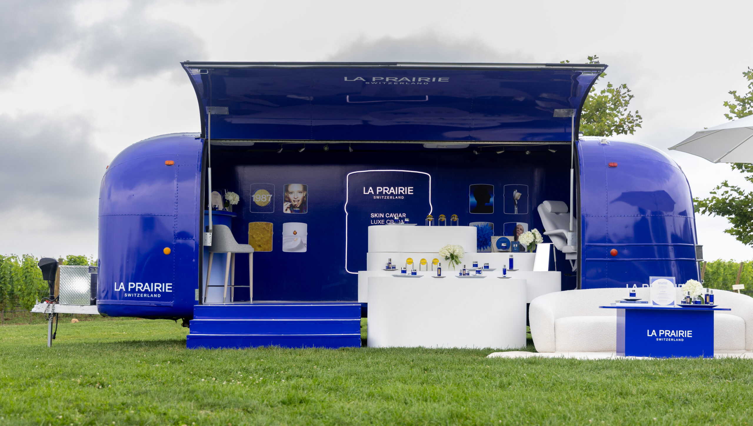 Blue La Prairie pop-up trailer with skincare products on white tables, outdoors on grass with trees.