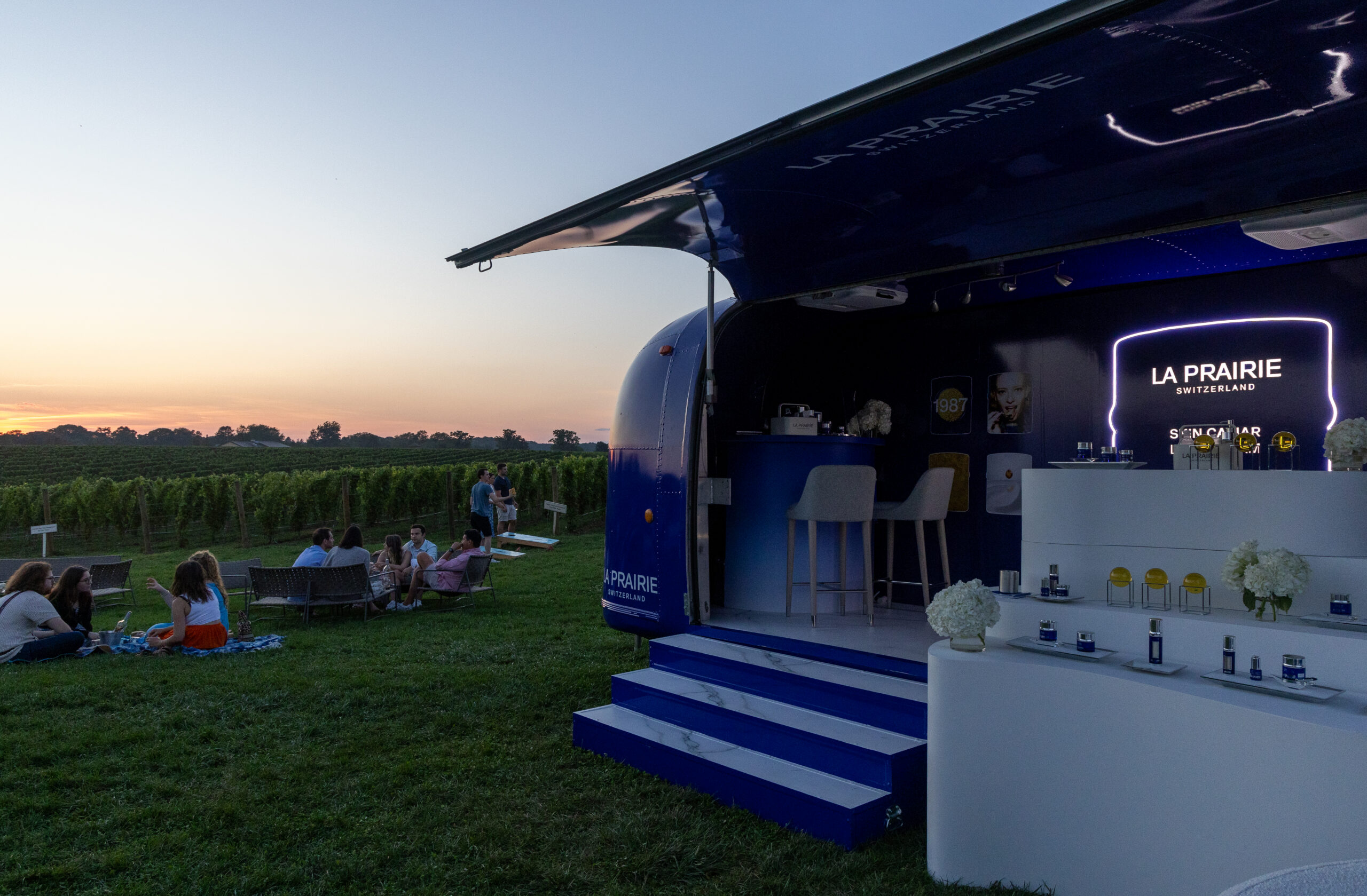 La Prairie pop up shop on grass at sunset with people relaxing near a vineyard, skincare on display.
