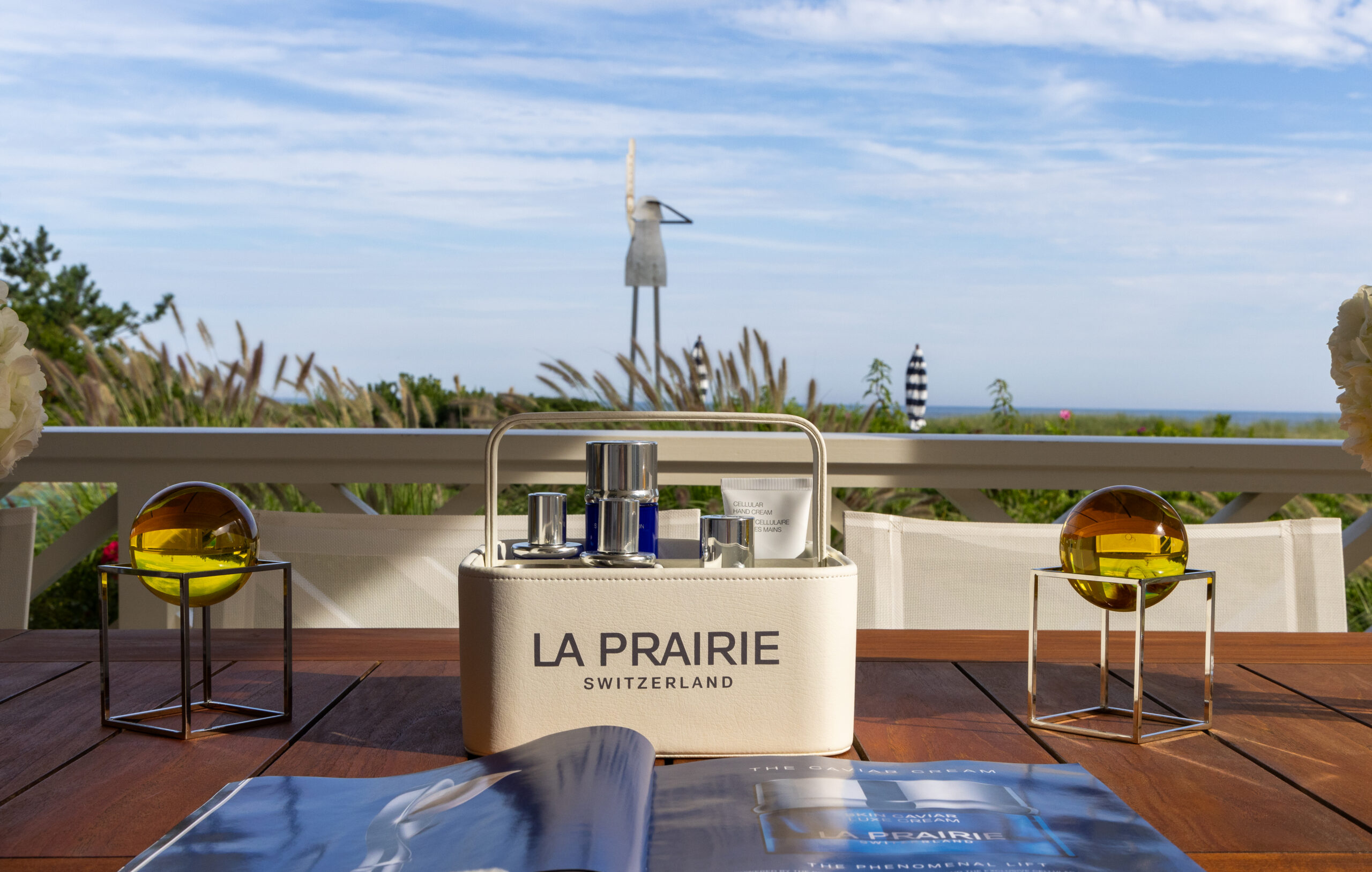 La Prairie skincare set on a wooden table with glass orbs, greenery, sculpture, and ocean behind.