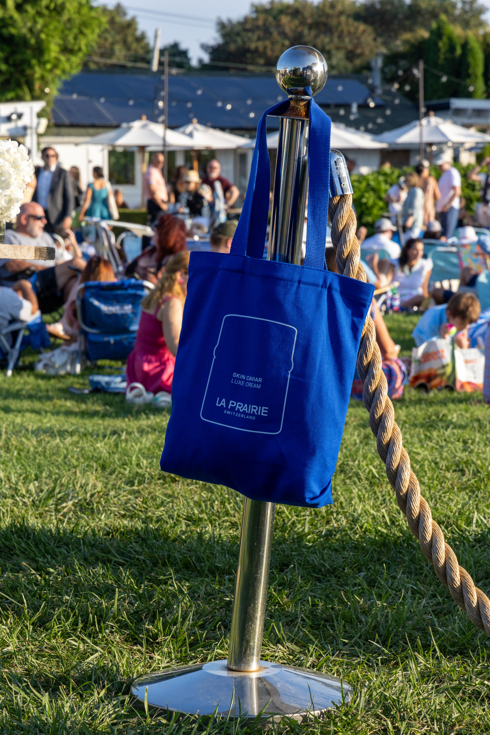 Blue tote bag with white text hangs on a stanchion post at La Prairie pop-up on green grass.
