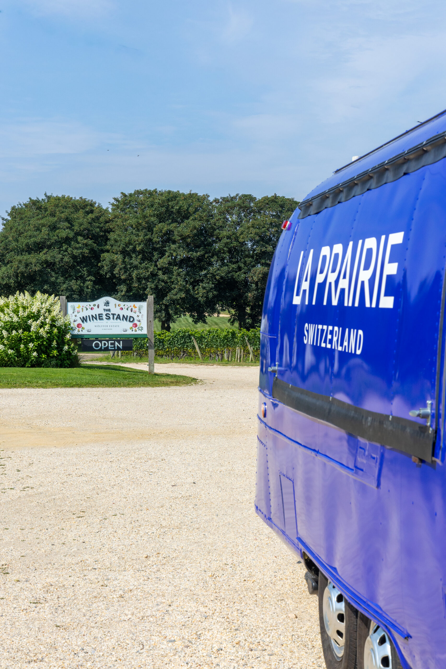 Blue LA PRAIRIE SWITZERLAND food truck parked on gravel; Wine Stand sign and trees in back.